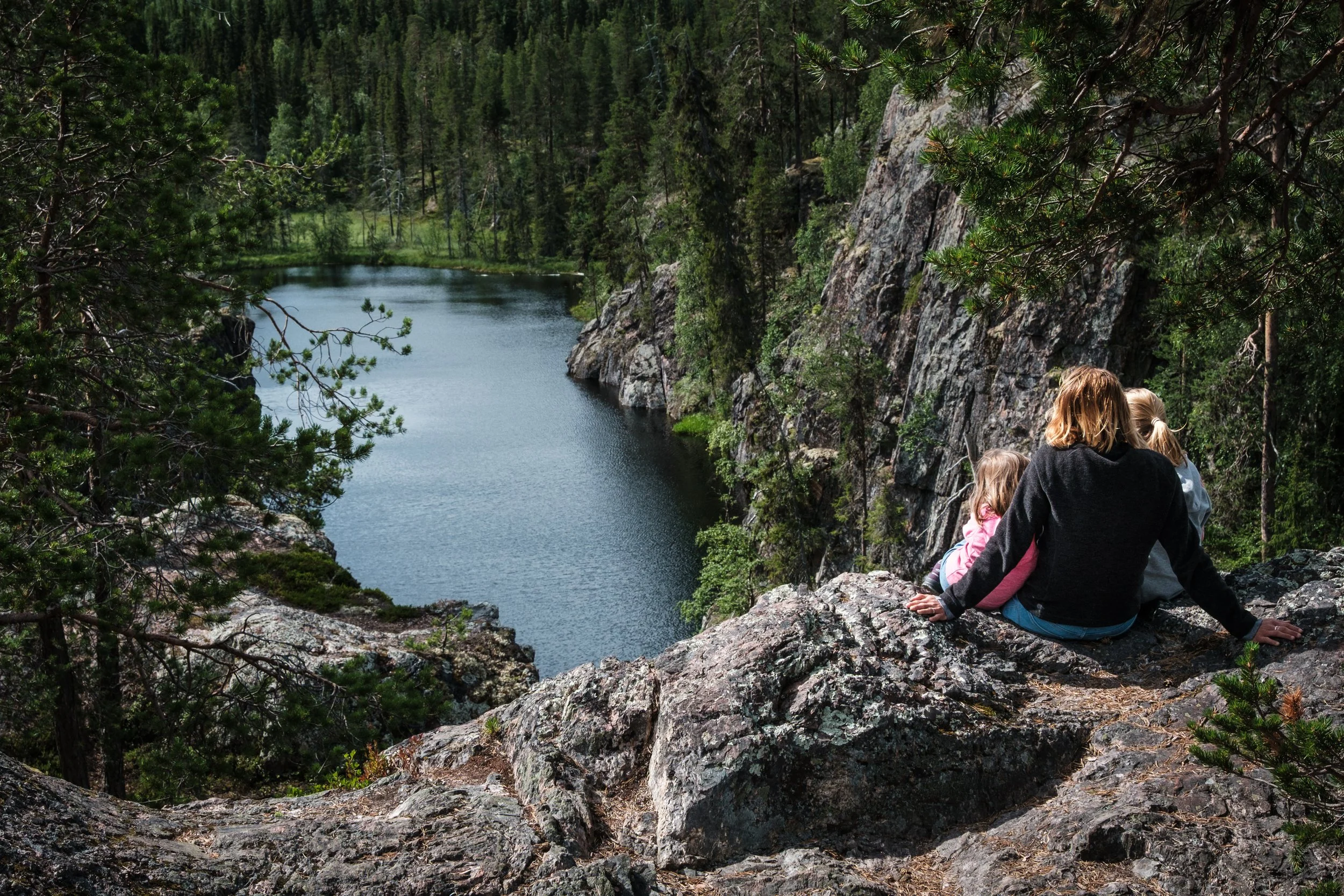 Drei Kinder sitzen auf einem Felsen am Rand eines Sees, umgeben von Bäumen und Felsen, in einer Berglandschaft.