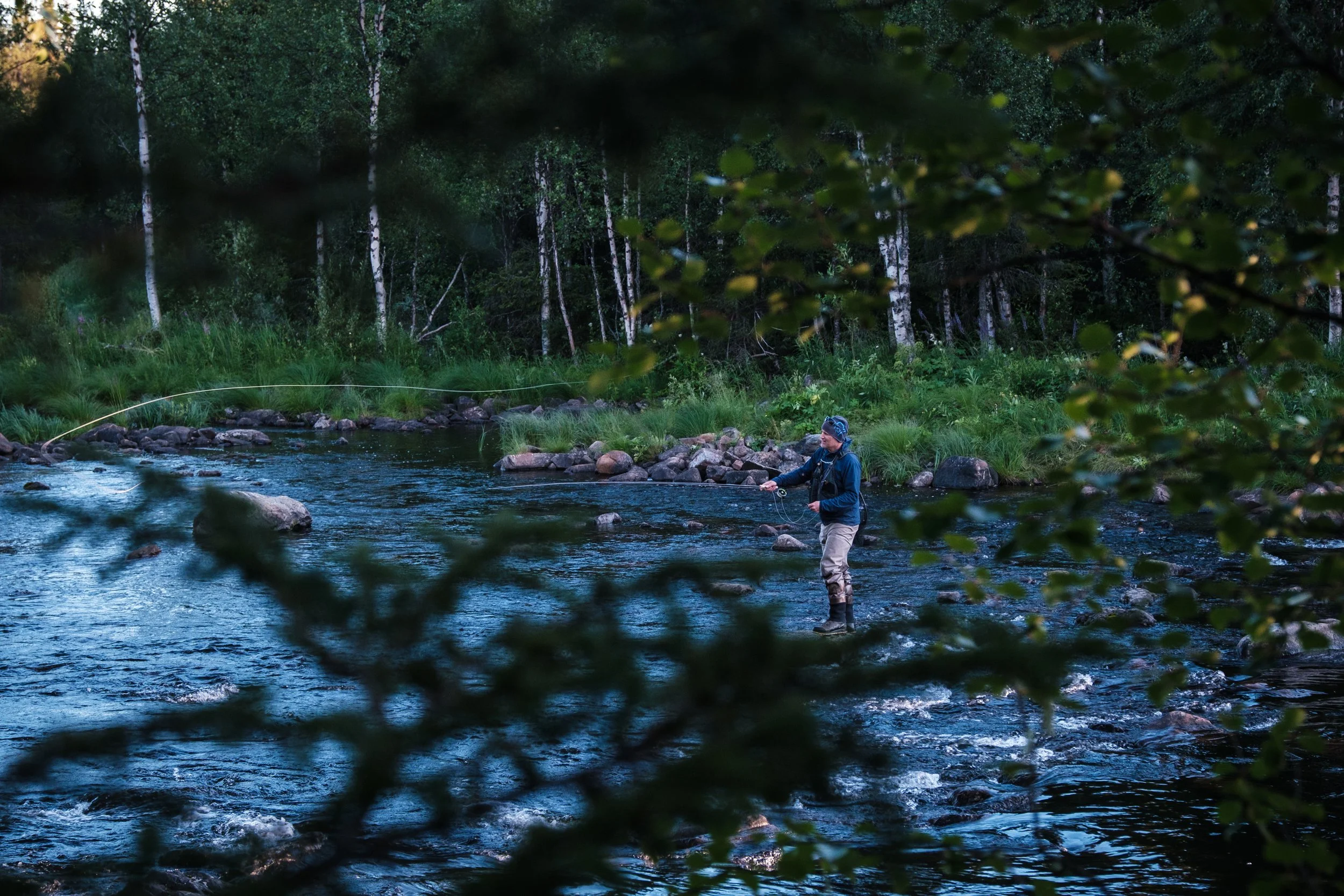 Ein Fischer steht in einem Fluss, umgeben von Bäumen und grüner Vegetation, und fischt bei Dämmerung.