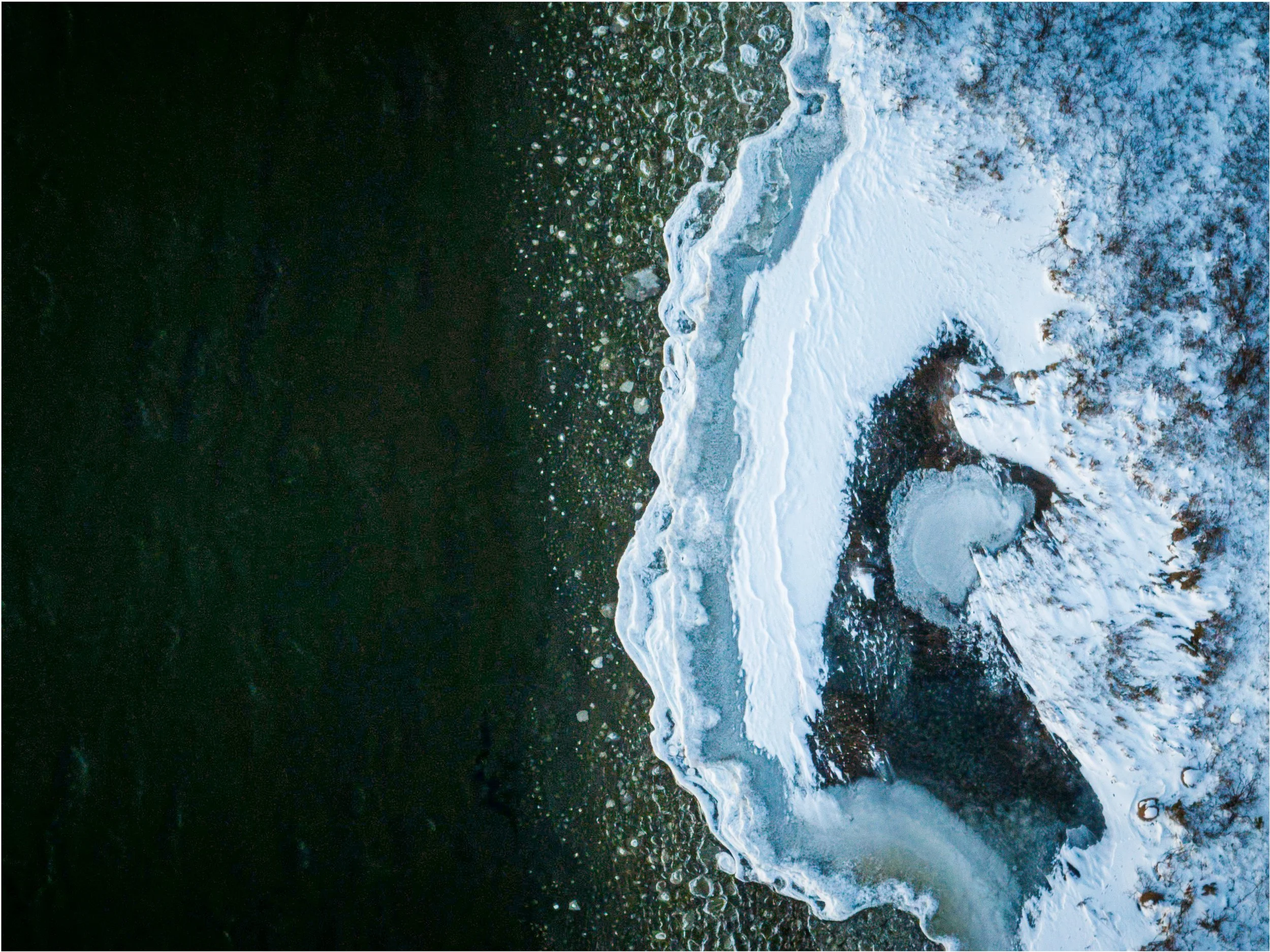 Vogelperspektive auf gefrorenen Fluss, der sich durch eine verschneite Landschaft zieht, mit Eisformationen und Bäumen am Ufer im Hintergrund.