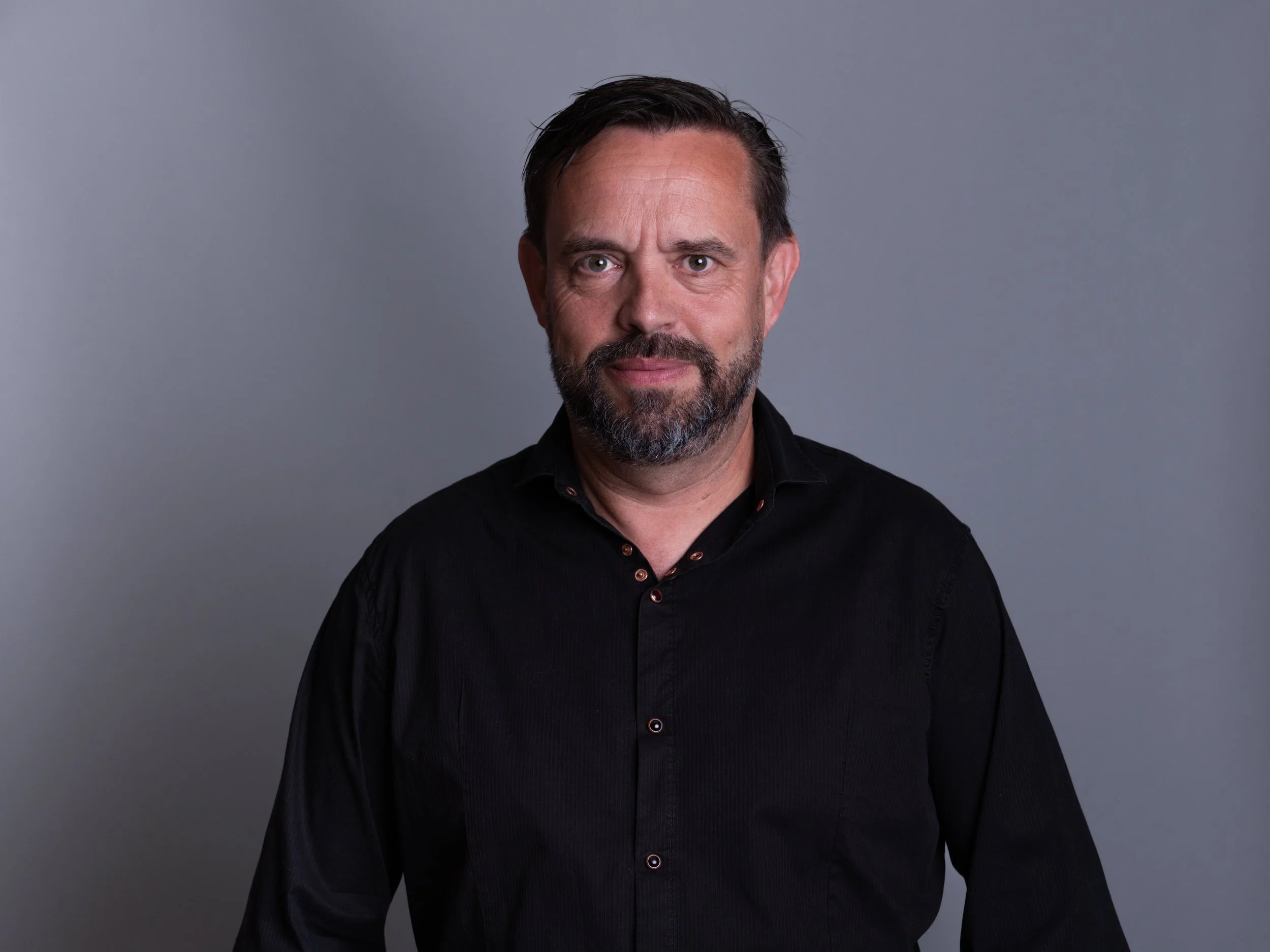 Ein Mann mit dunklem Haar, Bart und schwarzem Hemd vor grauem Hintergrund.