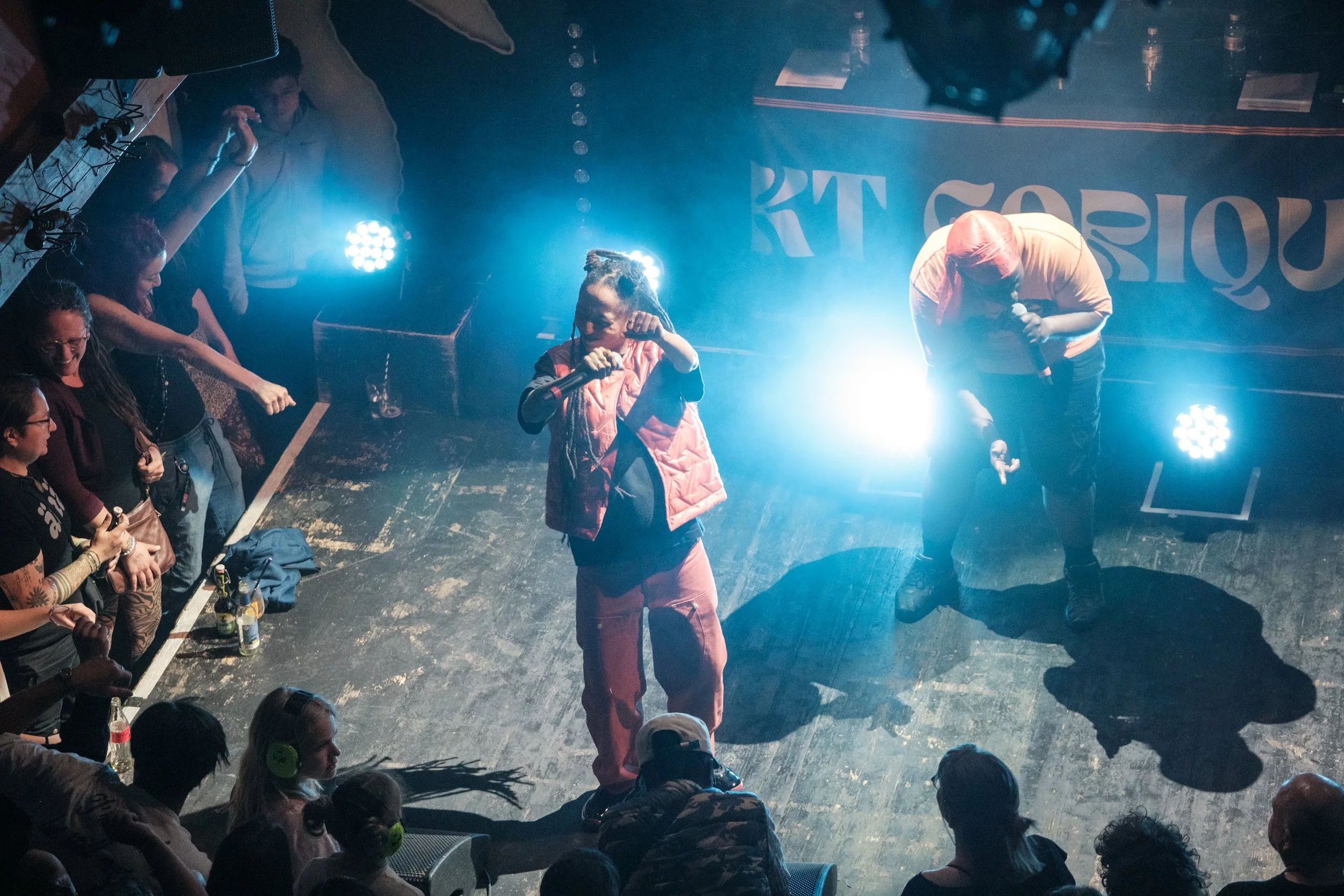 Zwei Performer auf einer Bühne, die in Mikrofone singen, eingerahmt von einem Publikum, das applaudiert und Fotos macht, bei einer Live-Performance in einem Club, mit bunten Lichteffekten und einem dunklen Holzparkettboden.