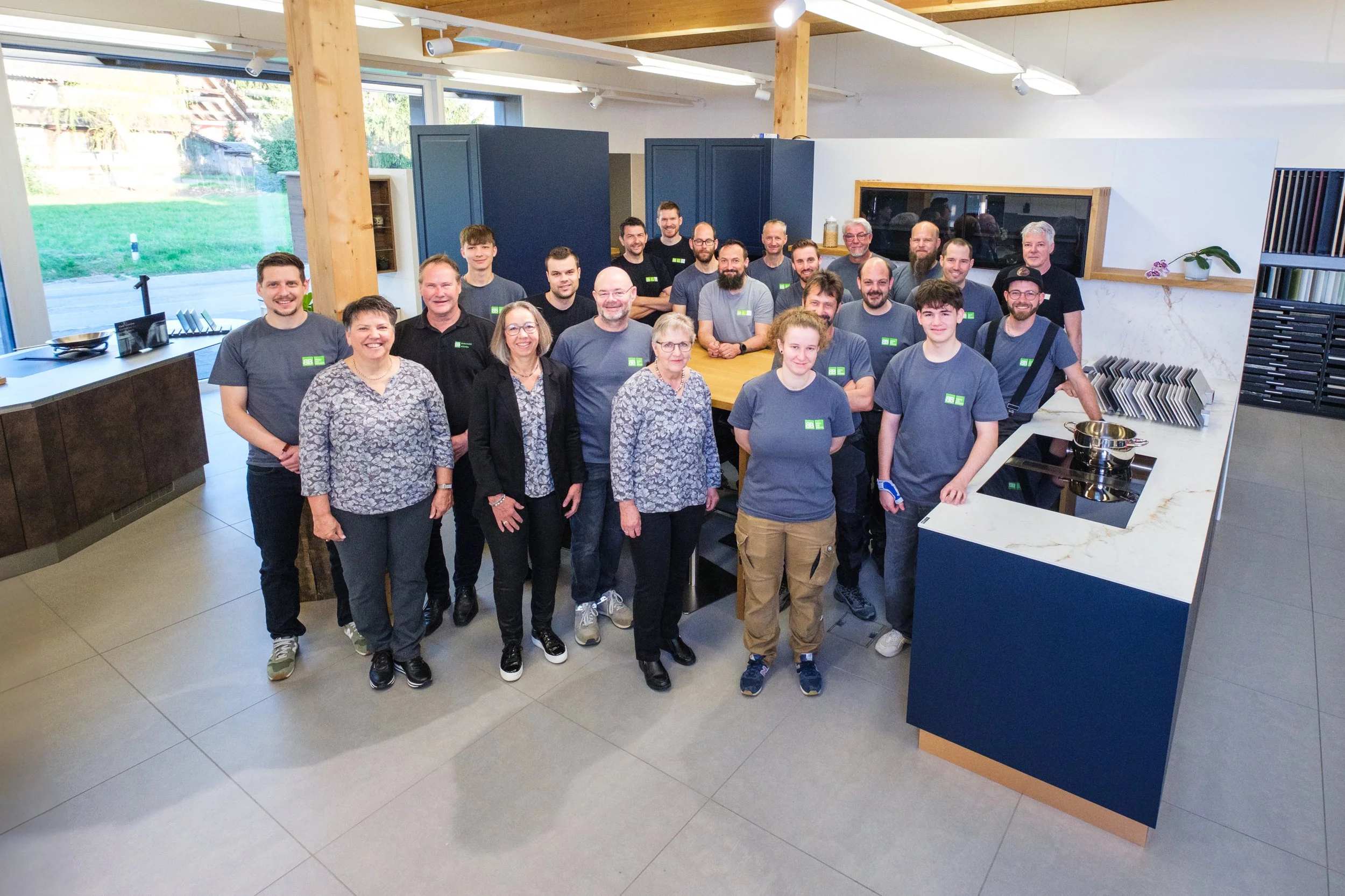 Gruppe von Menschen in einer modernen Küchenumgebung, einige in grauen T-Shirts, vor einem weißen Arbeitsbereich mit Kochutensilien und Dekorationen.