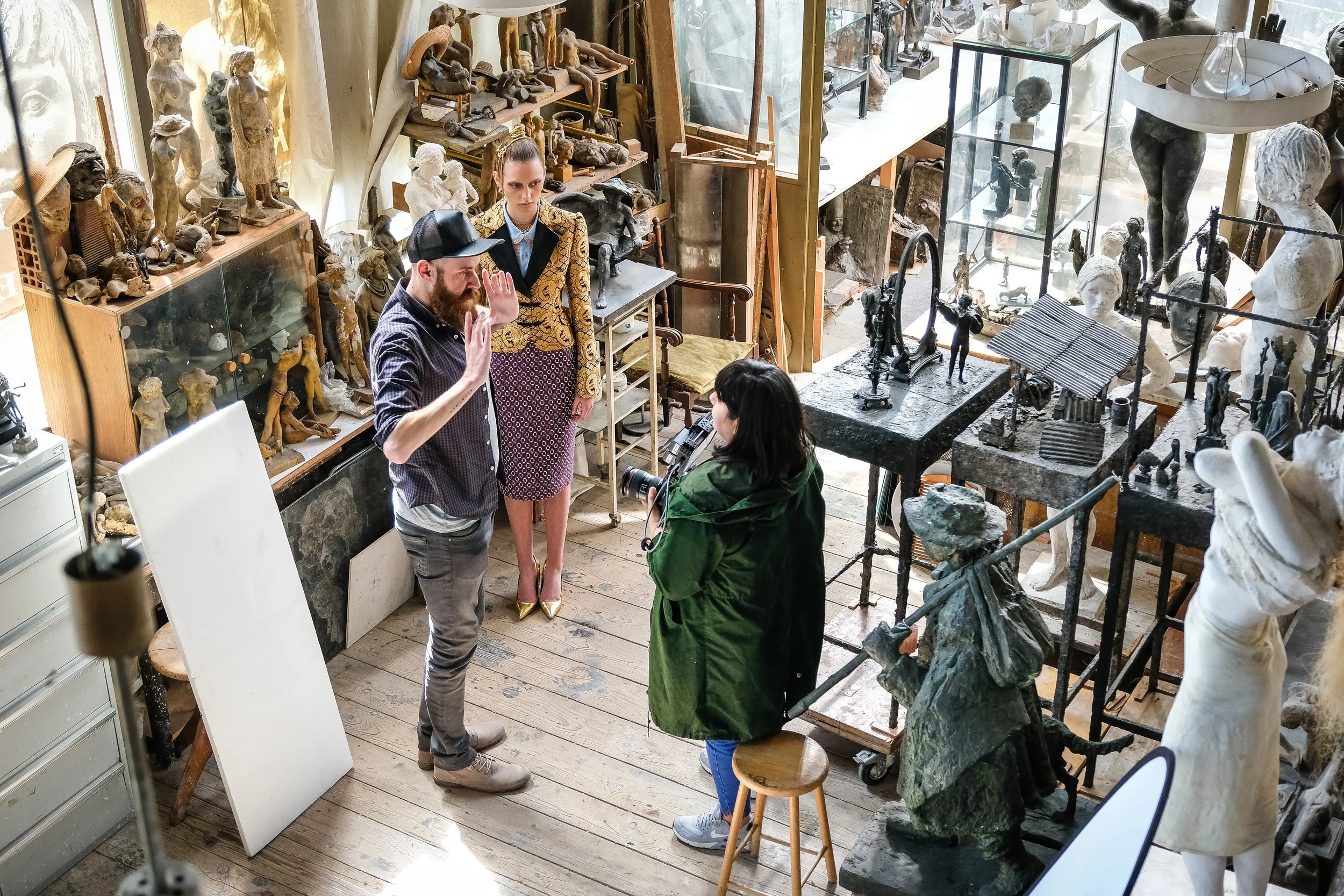 Drei Menschen in einem Antiquitätengeschäft, darunter ein Mann, der eine Handbewegung macht, eine Frau, die zuhört, und eine weitere Frau, die mit einer Kamera filmt, umgeben von Skulpturen und Antiquitäten.