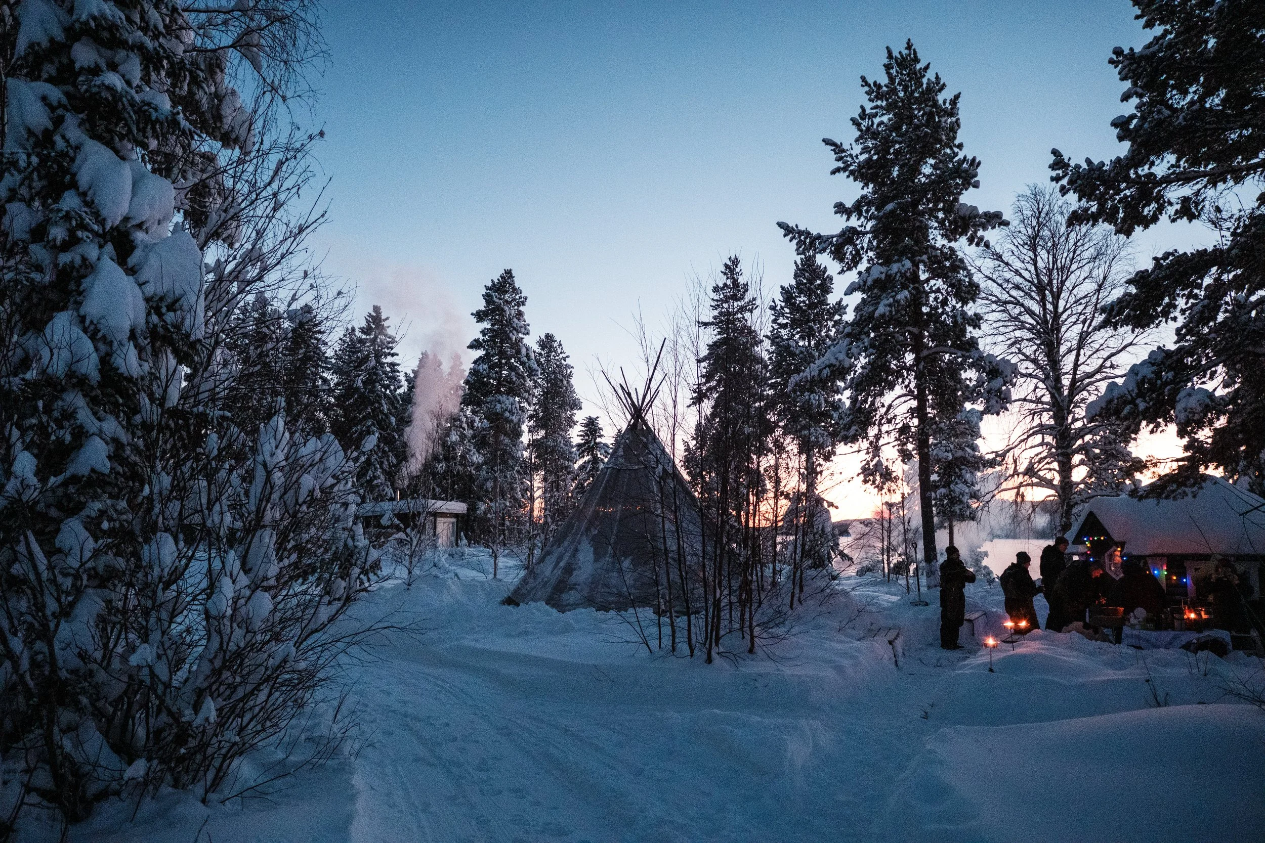 Schneebedeuter Wald bei Sonnenuntergang mit Zelten und Menschen, die um Feuer stehen.