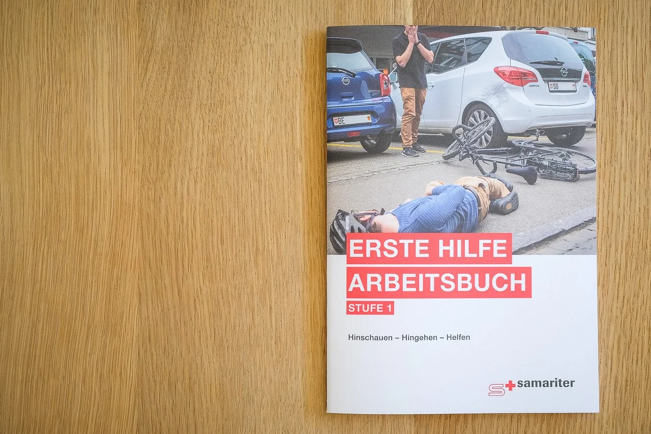 Lehrbuch für Erste Hilfe am Arbeitsplatz mit dem Titel "ERSTE HILFE ARBEITSBUCH STUFE 1" auf einem Holztisch. Das Cover zeigt eine Szene mit einem verletzten Fahrradfahrer und einem anderen Menschen, der bei einem Unfall hilft.