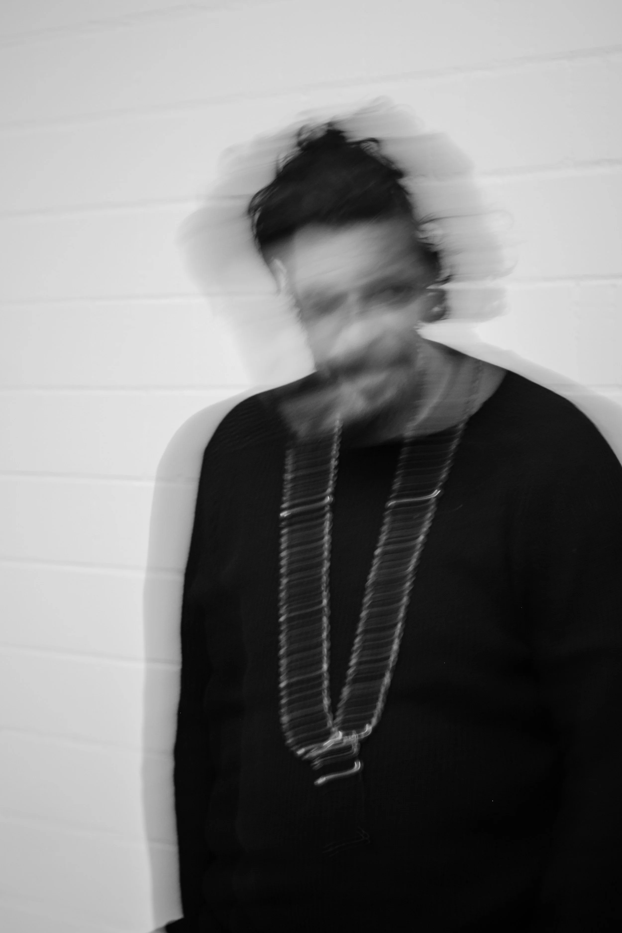 Schwarz-weiß-Foto eines Mannes mit dunklen, zerzausten Haaren und Bart, der vor einer weißen Wand steht. Der Mann trägt eine dunkle Jacke und eine lange Kette.