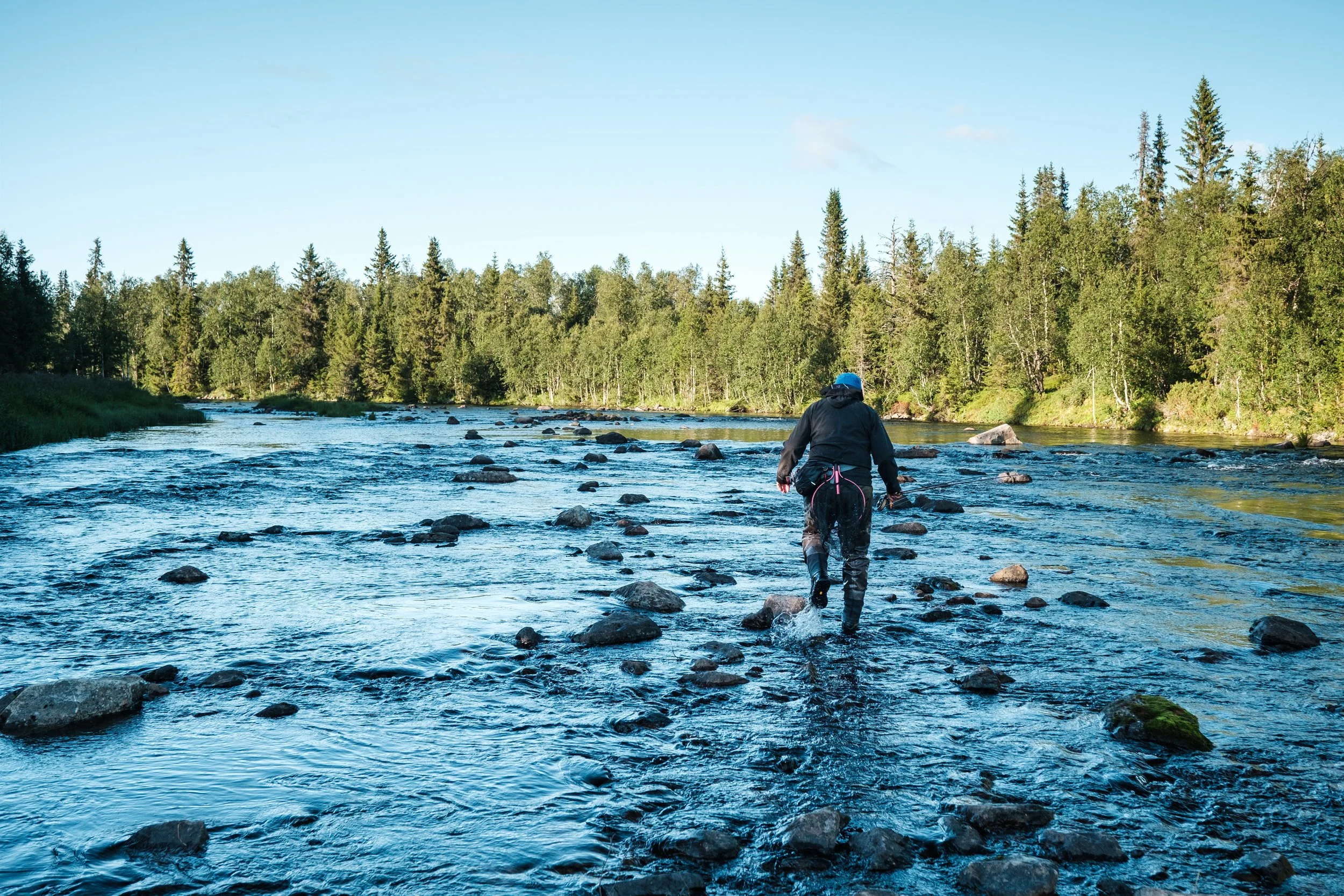 Person in dunkler Kleidung und blauer Mütze wandert durch einen flachen Fluss in einer bewaldeten Umgebung.