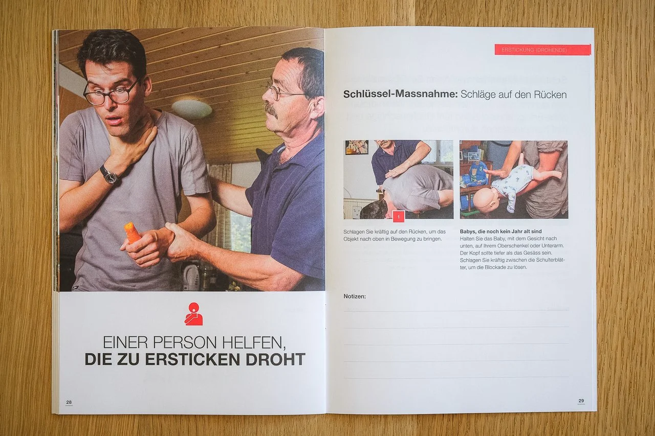 Zwei Männer, wobei einer einem anderen die Hand auf den Rücken legt, während dieser mit der Hand an der Kehle hält. Das Bild befindet sich in einem Ratgeberbuch über Erste Hilfe bei Erstickungsgefahr.