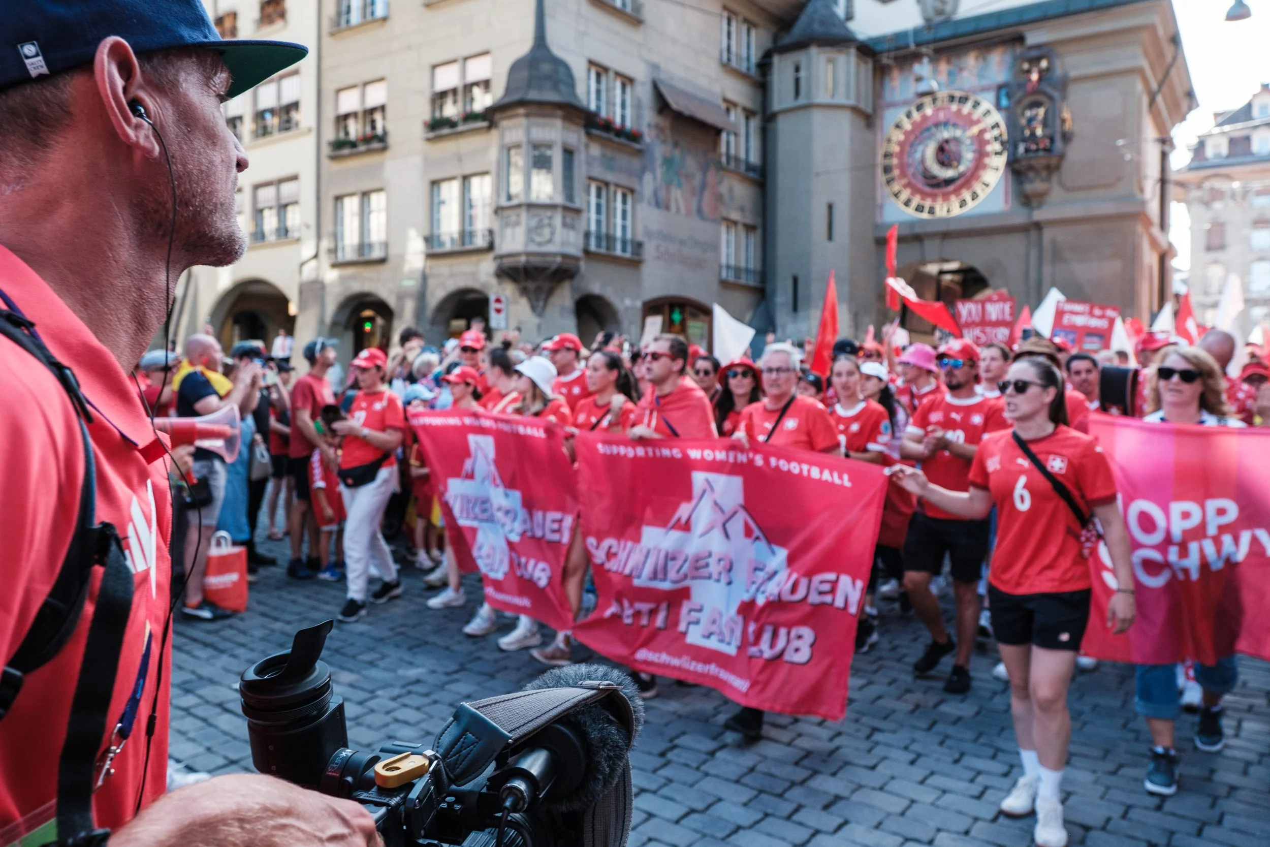 Gruppe von Frauen und Männern, die bei einer Demonstration in roten T-Shirts und Hüten eine große rote Fahne mit weißen und pinken Texten halten, im Hintergrund historische Gebäude und eine Uhr