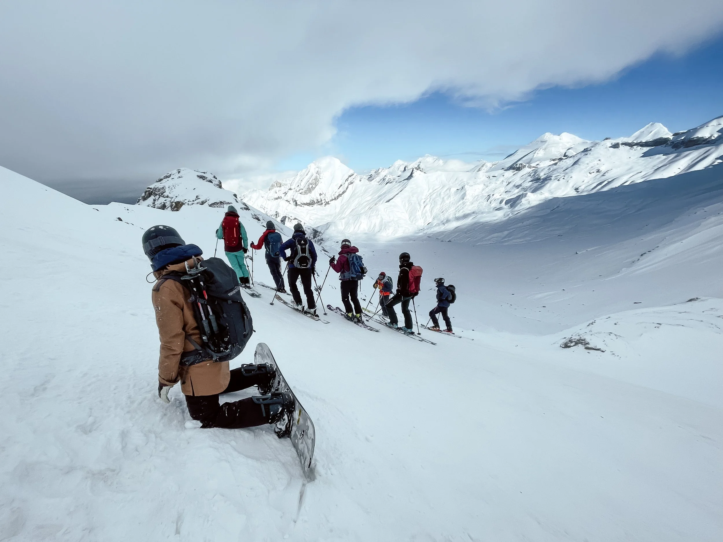 Gruppe von Skifahrern in Winterkleidung auf einer Schneefläche in den Bergen, eine Person steht mit Schneeschuhen im Vordergrund, die anderen fahren in einer Reihe weiter oben im Schnee.