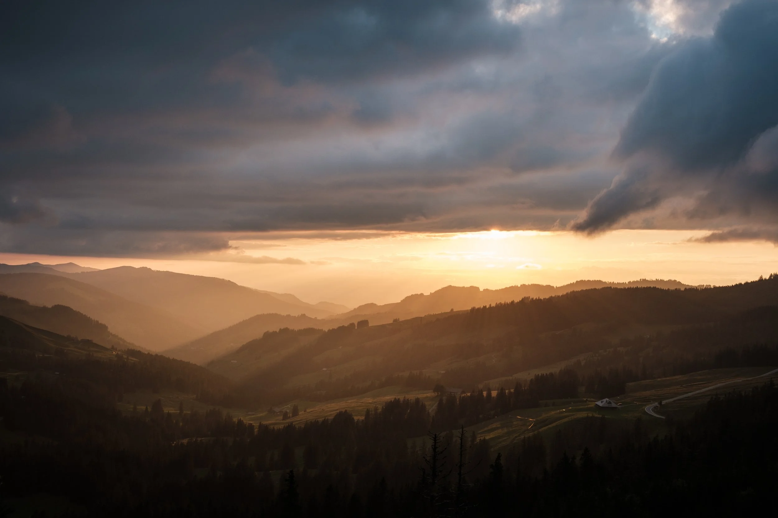 Berglandschaft bei Sonnenuntergang mit dunklen Wolken am Himmel und fahlem Licht auf den Hügeln im Vordergrund.