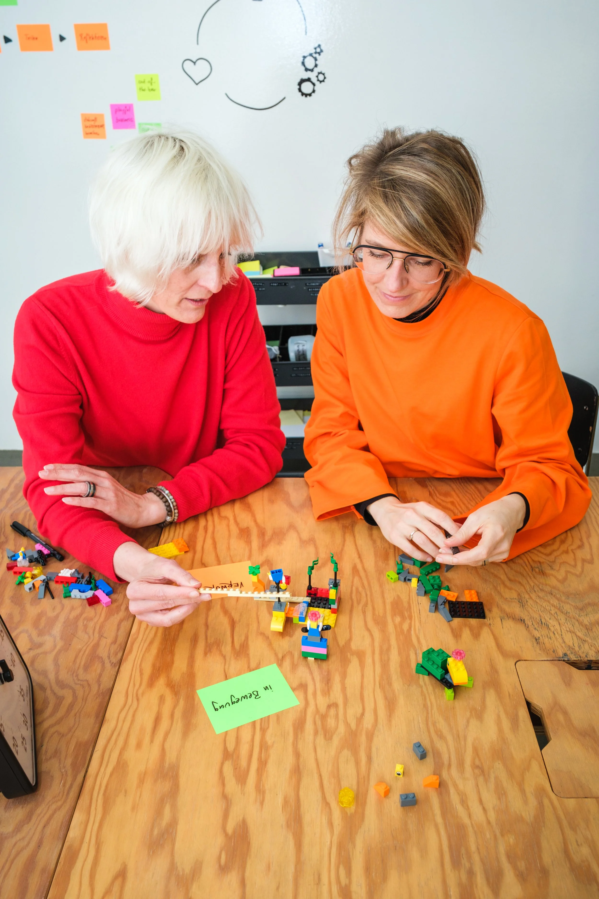 Zwei Frauen sitzen an einem Tisch und bauen mit LEGO-Steinen. Im Hintergrund sind ein Whiteboard und eine Wand mit Post-it-Notizen und einer einfachen Zeichnung eines Gesichts mit Herz, Kreisscheiben und Blumen. Die Frauen tragen rote und orange Klei