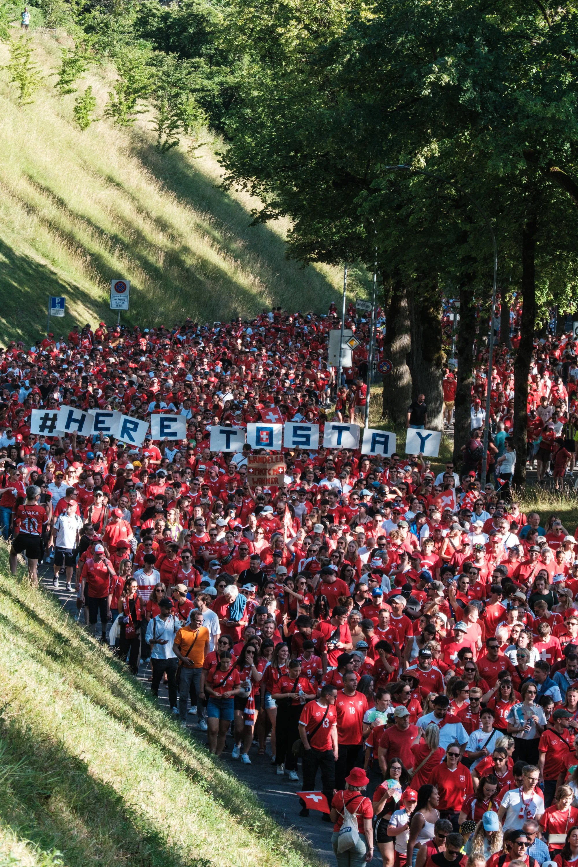 Eine große Menschenmenge, die an einem sonnigen Tag in roten Trikots spazieren geht. In der Mitte halten sie Schilder mit der Aufschrift "#HERESTAY" und es sind viele schweizer Flaggen sichtbar, was auf eine Unterstützung für die Schweiz hindeutet.