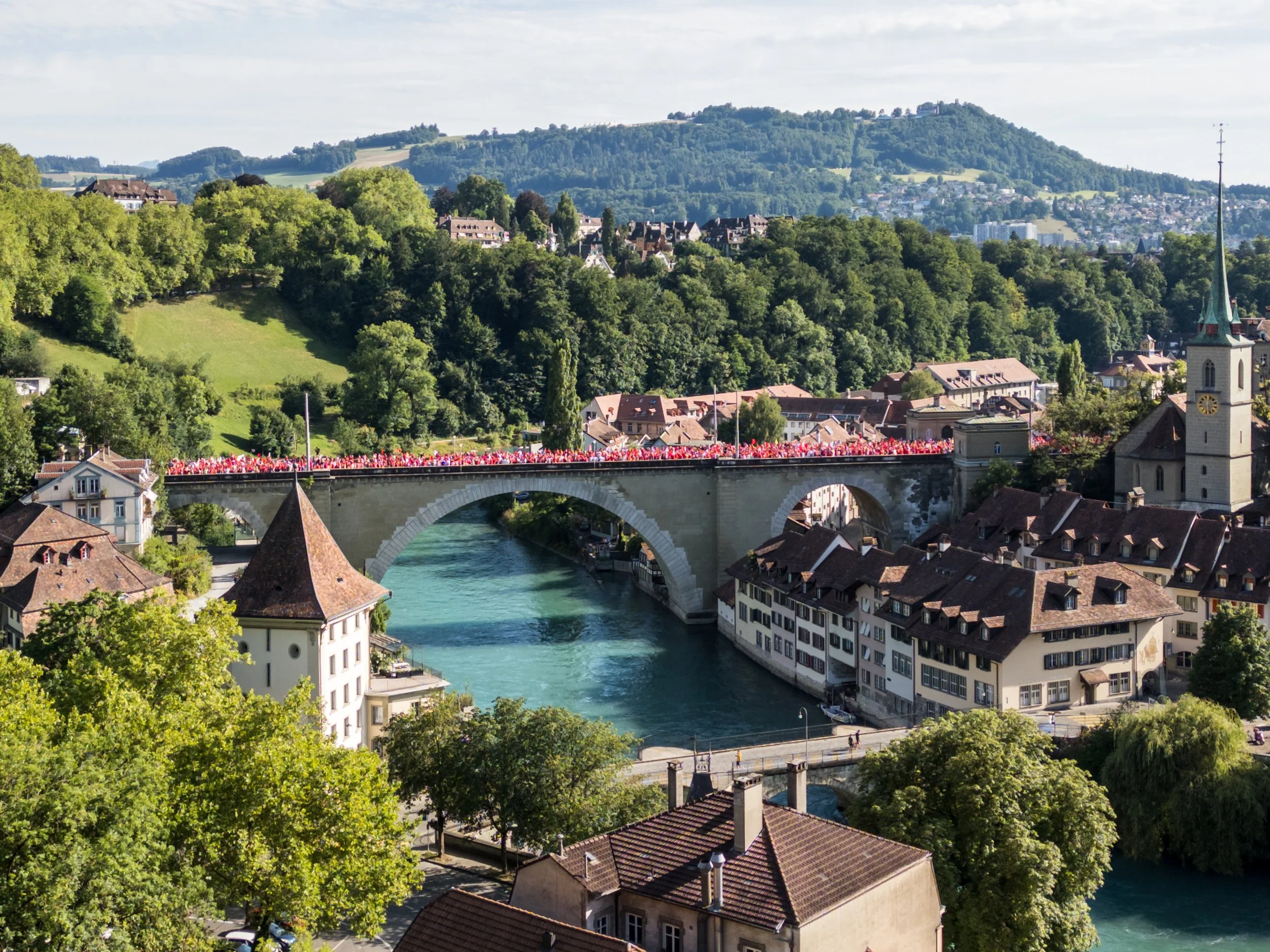 Blick auf eine Brücke über einen Fluss in einer mittelalterlichen Stadt, umgeben von grünen Hügeln und Bergen, mit einigen Gebäuden und einer Kirche mit Turm.