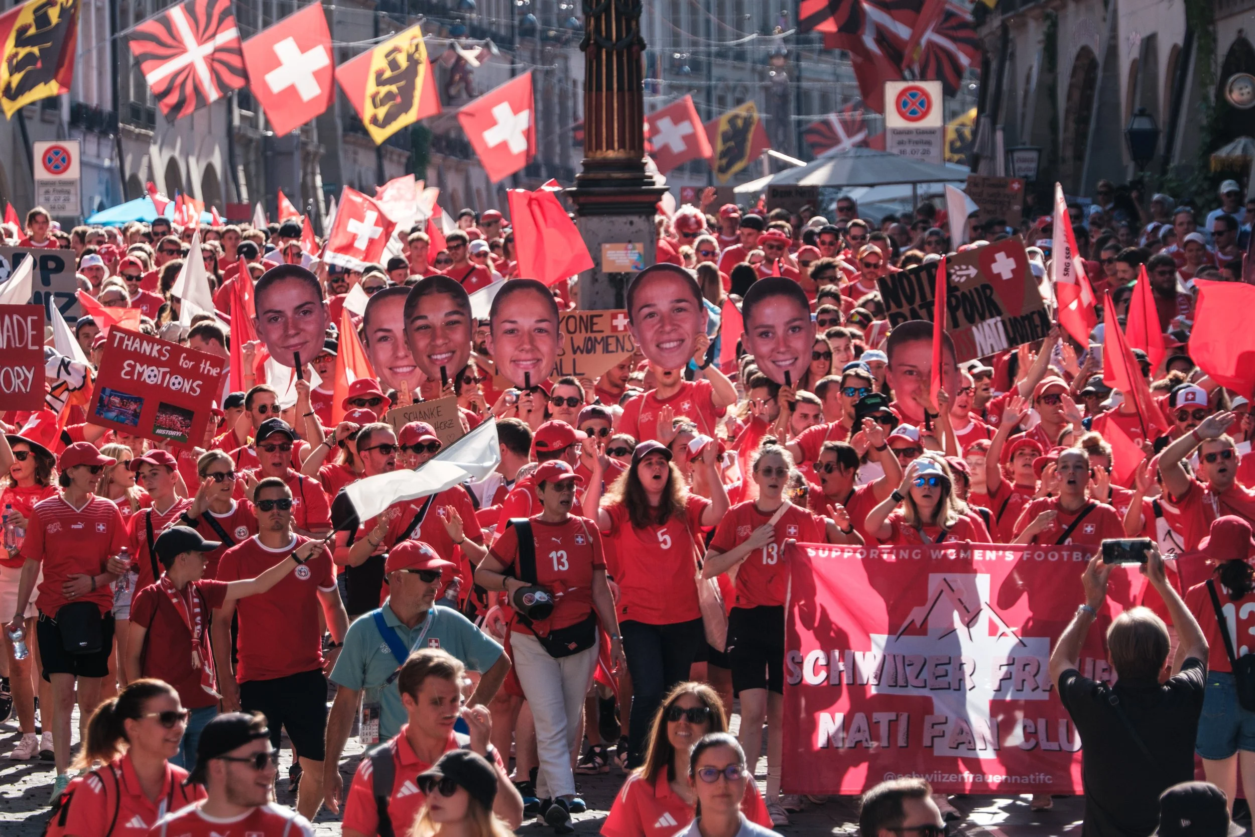 Viele Menschen in roter Kleidung bei einer Demonstration oder einem Fest, die Schweizer Fahnen und große Fotos von Frauenagierten tragen.