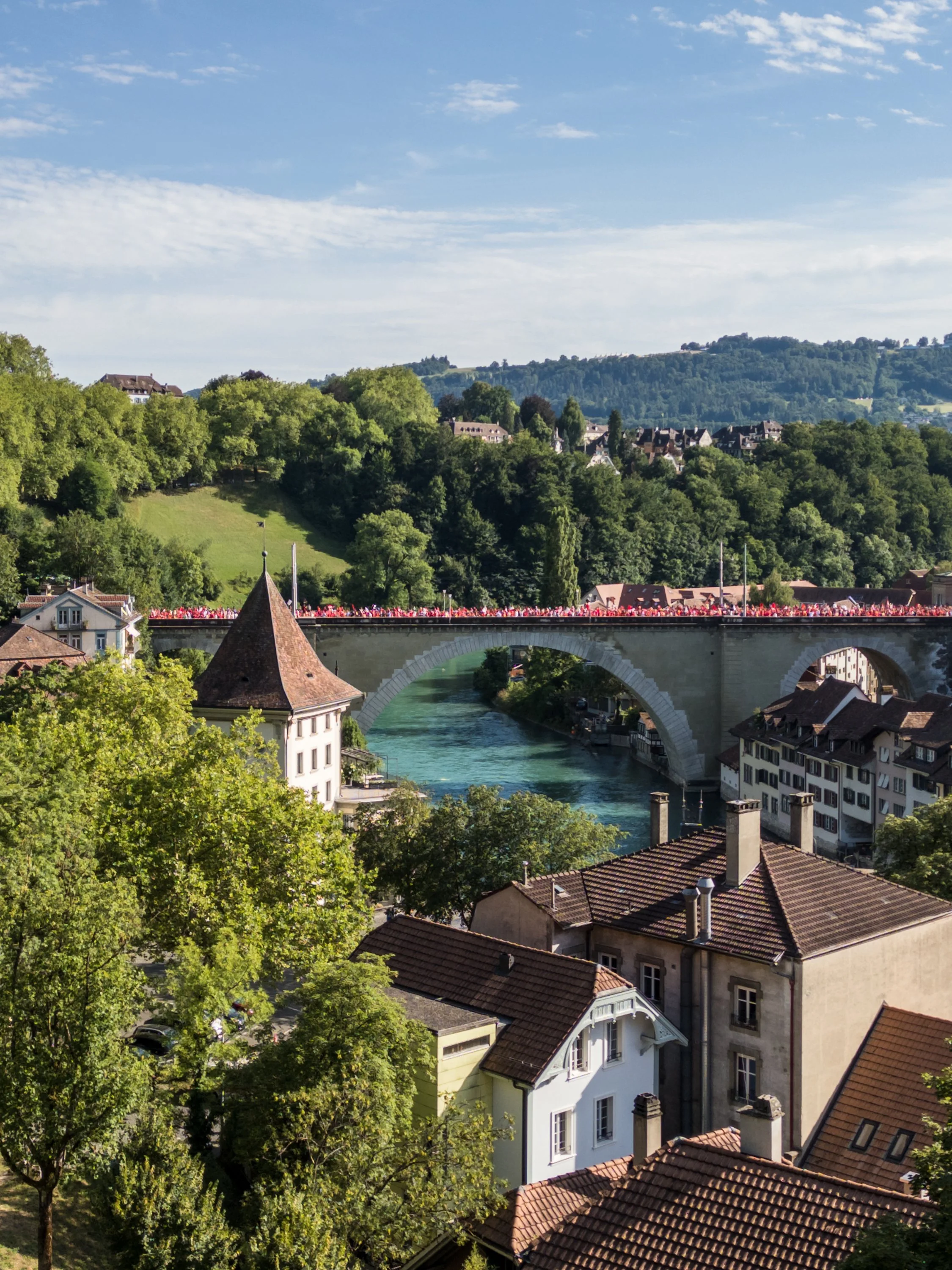 Eine Stadt mit traditionellen Häusern und einer Brücke über einen Fluss, umgeben von grünen Hügeln bei sonnigem Wetter.