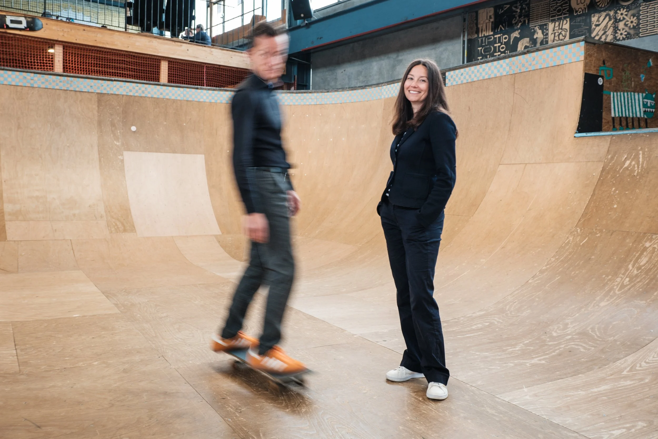 Zwei Personen, eine fahrend mit Skateboard, die andere stehenden, in einem Skatepark mit Holzrampe.