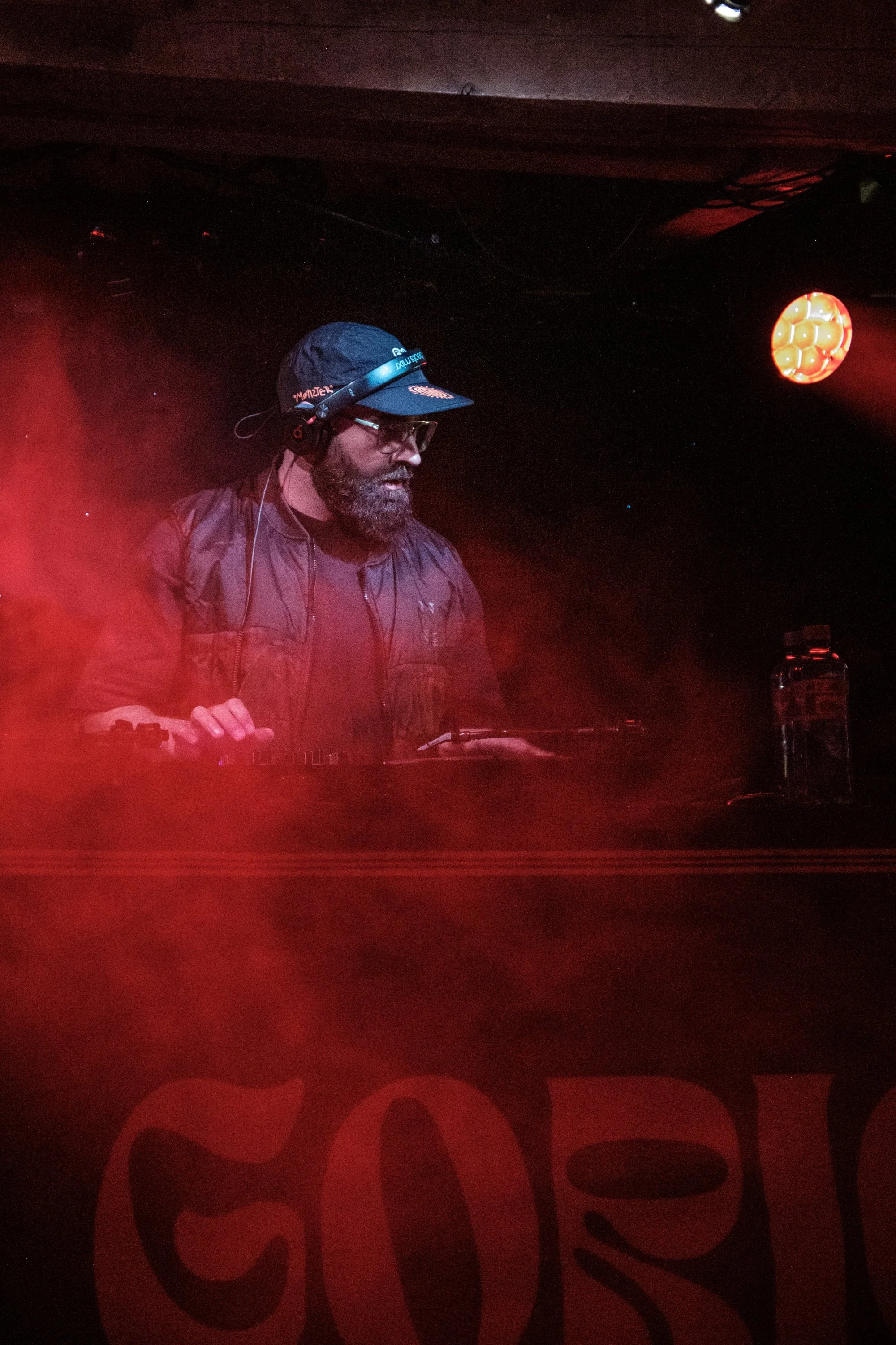 Ein DJ mit Hut und Brille hinter einem Mischpult, beleuchtet von rotem Licht in einem dunklen Club.