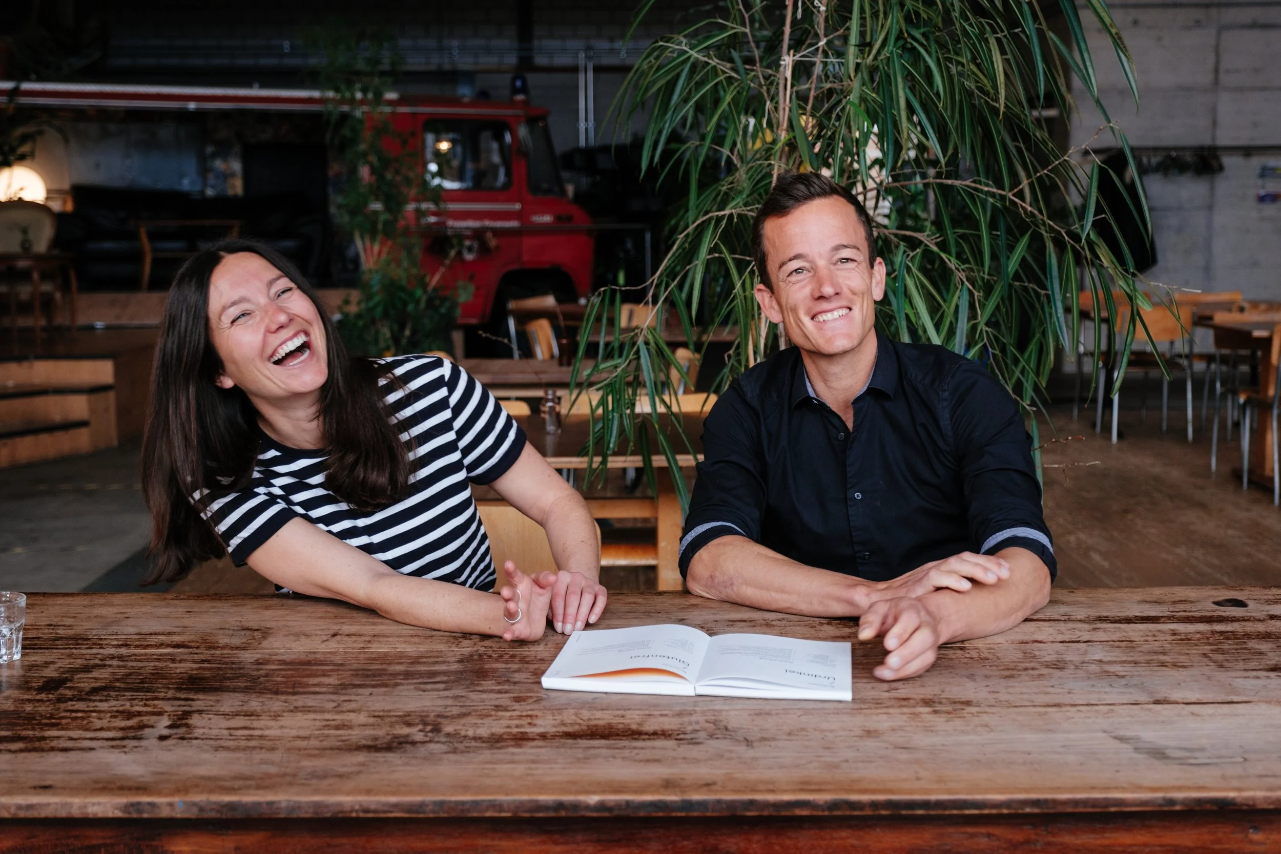 Ein Mann und eine Frau sitzen lachend an einem Holz Tisch in einem rustikalen Raum mit Pflanzen und einem roten Fahrzeug im Hintergrund.
