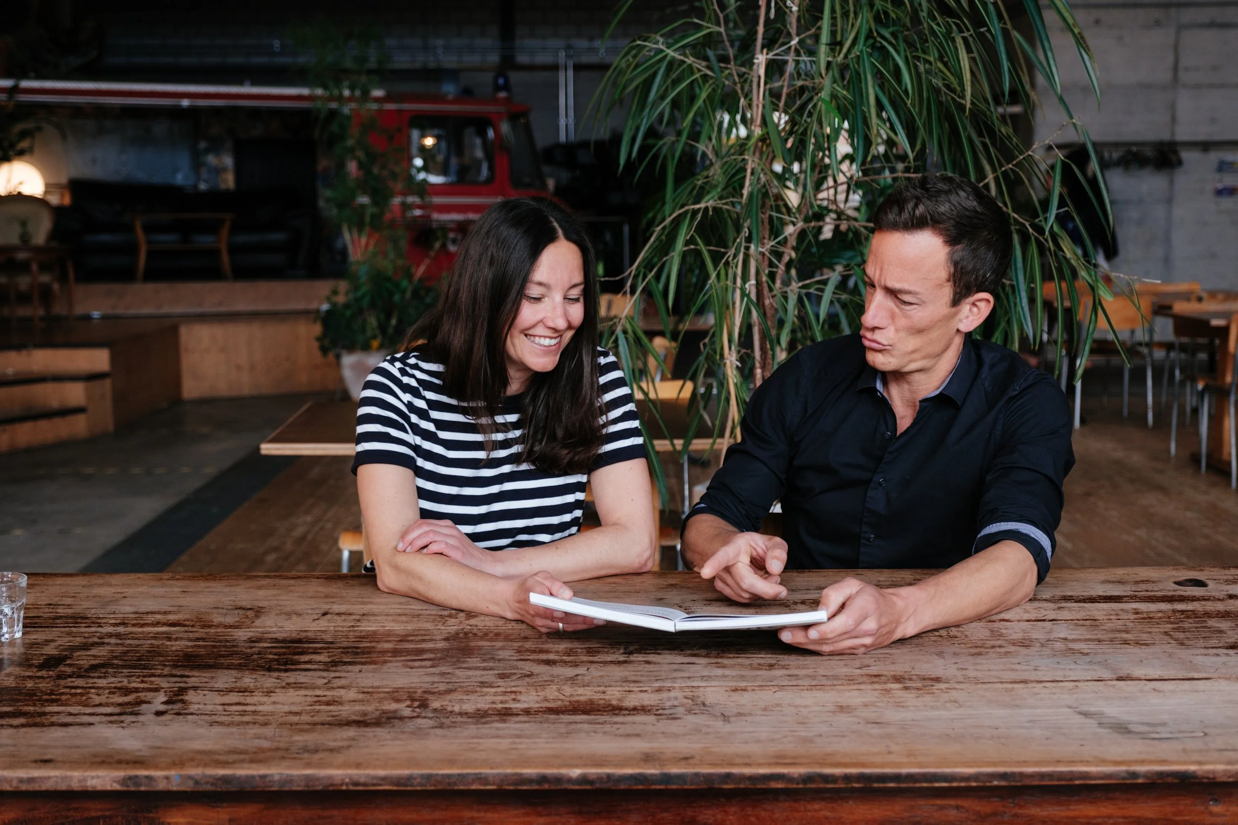 Zwei Menschen sitzen an einem Holztablett in einem Restaurant, schauen auf ein offenes Buch, lachen und diskutieren.