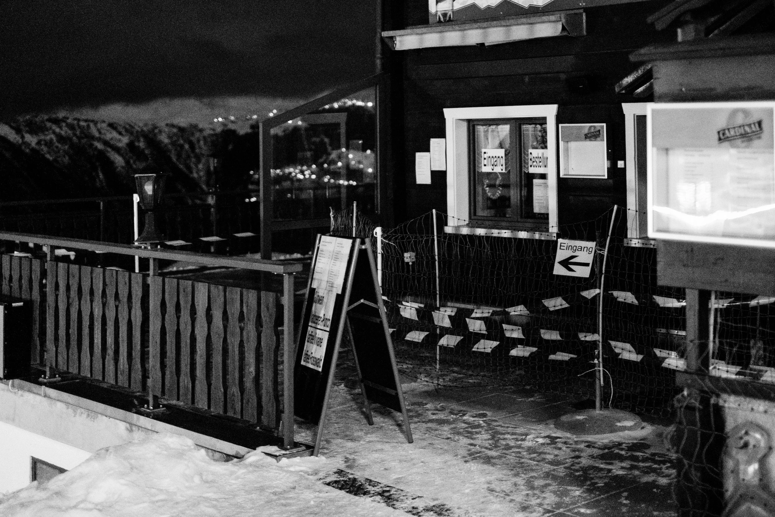 Schwarzes und weißes Bild eines Eingangsbereichs eines Gebäudes mit einem Fenster und mehreren Hinweisschildern, inklusive Schneefläche im Vordergrund. Es ist nachts, mit Bergen im Hintergrund.
