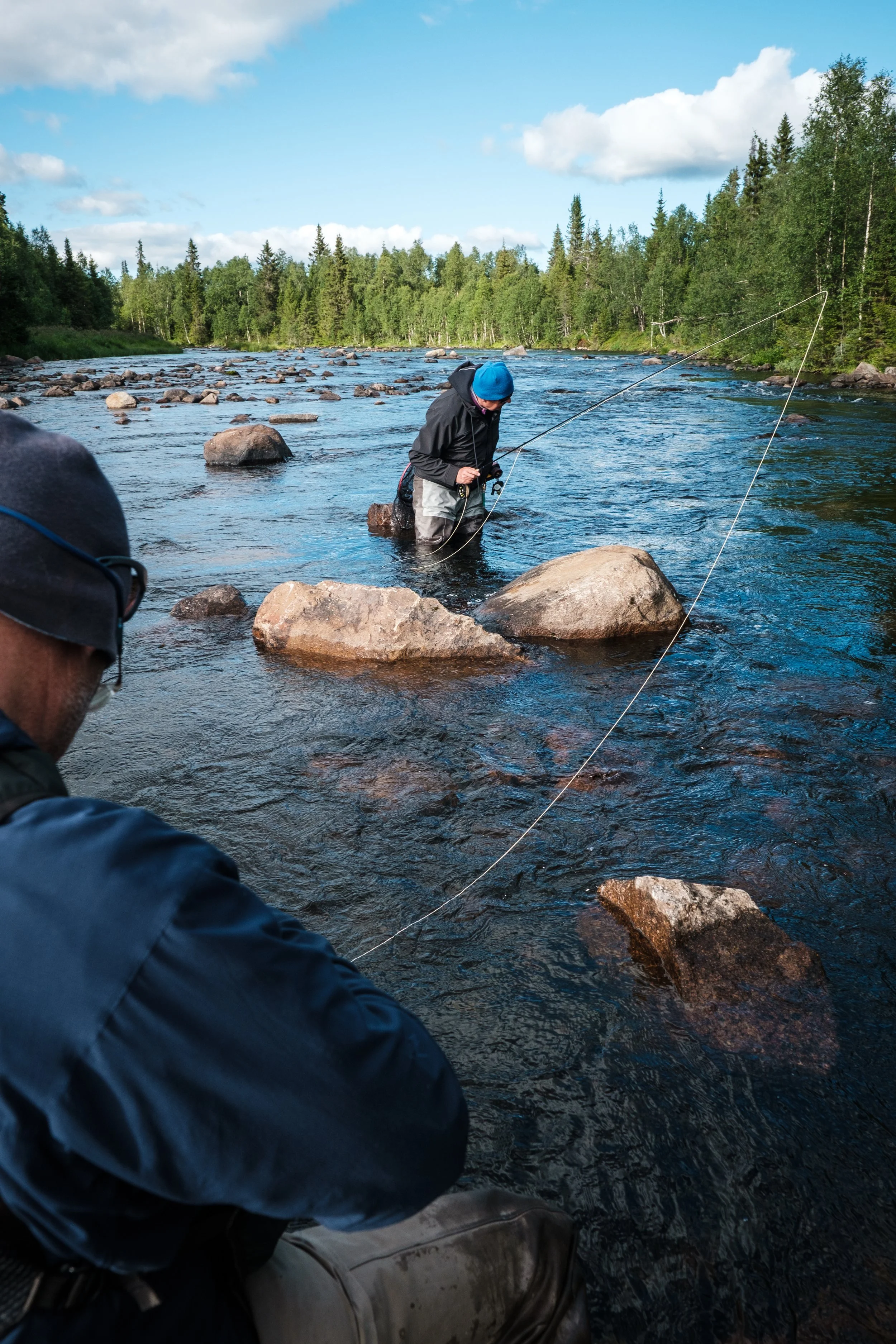 Zwei Männer, die in einem Fluss mit Steinen angeln, umgeben von Bäumen und blauer Himmel.