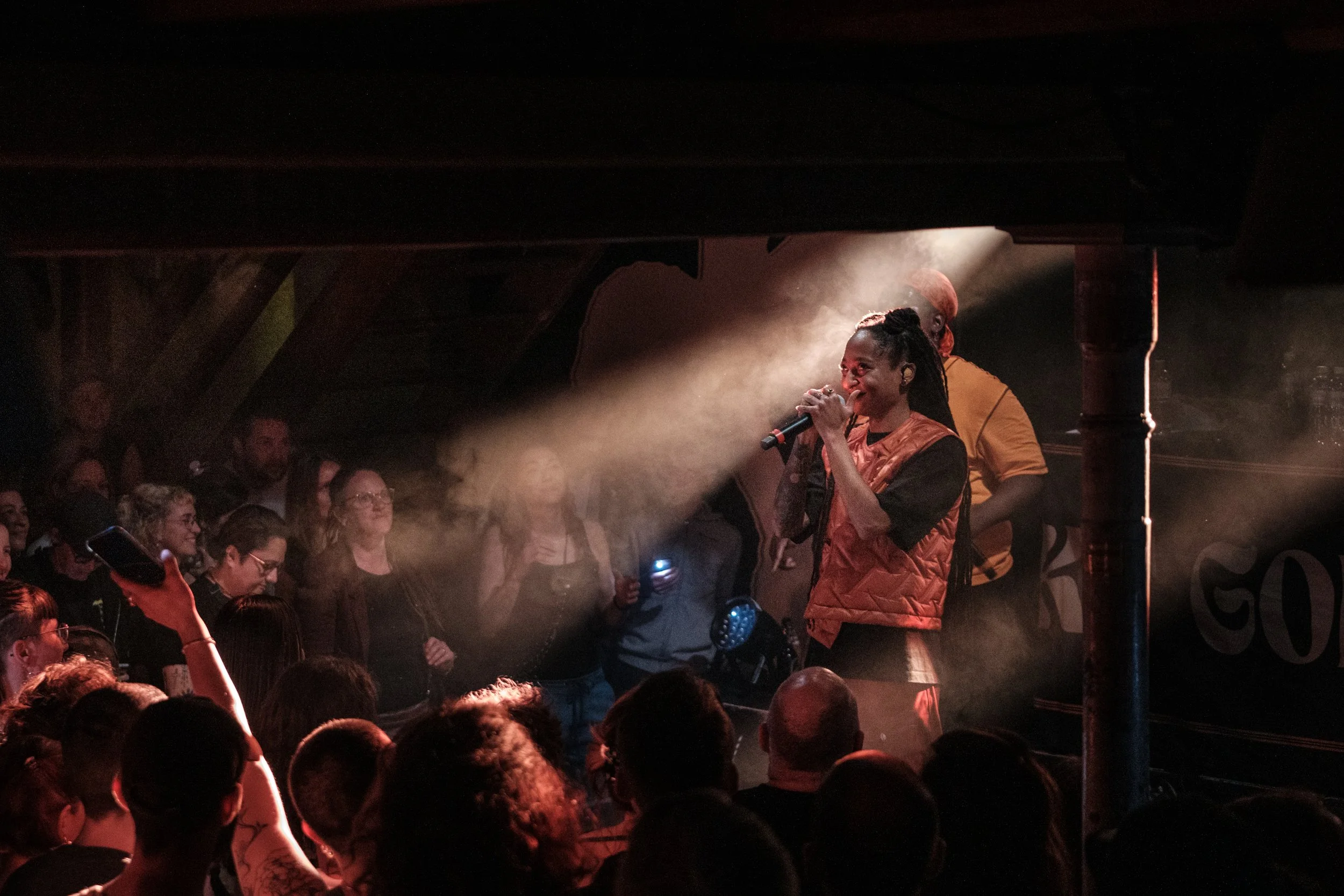 Eine Sängerin singt auf einer Bühne vor einem Publikum. Sie trägt eine orangefarbene Weste und hat Dreadlocks. Die Bühne ist mit Scheinwerfern beleuchtet und das Publikum sitzt oder steht in der dunklen Umgebung.