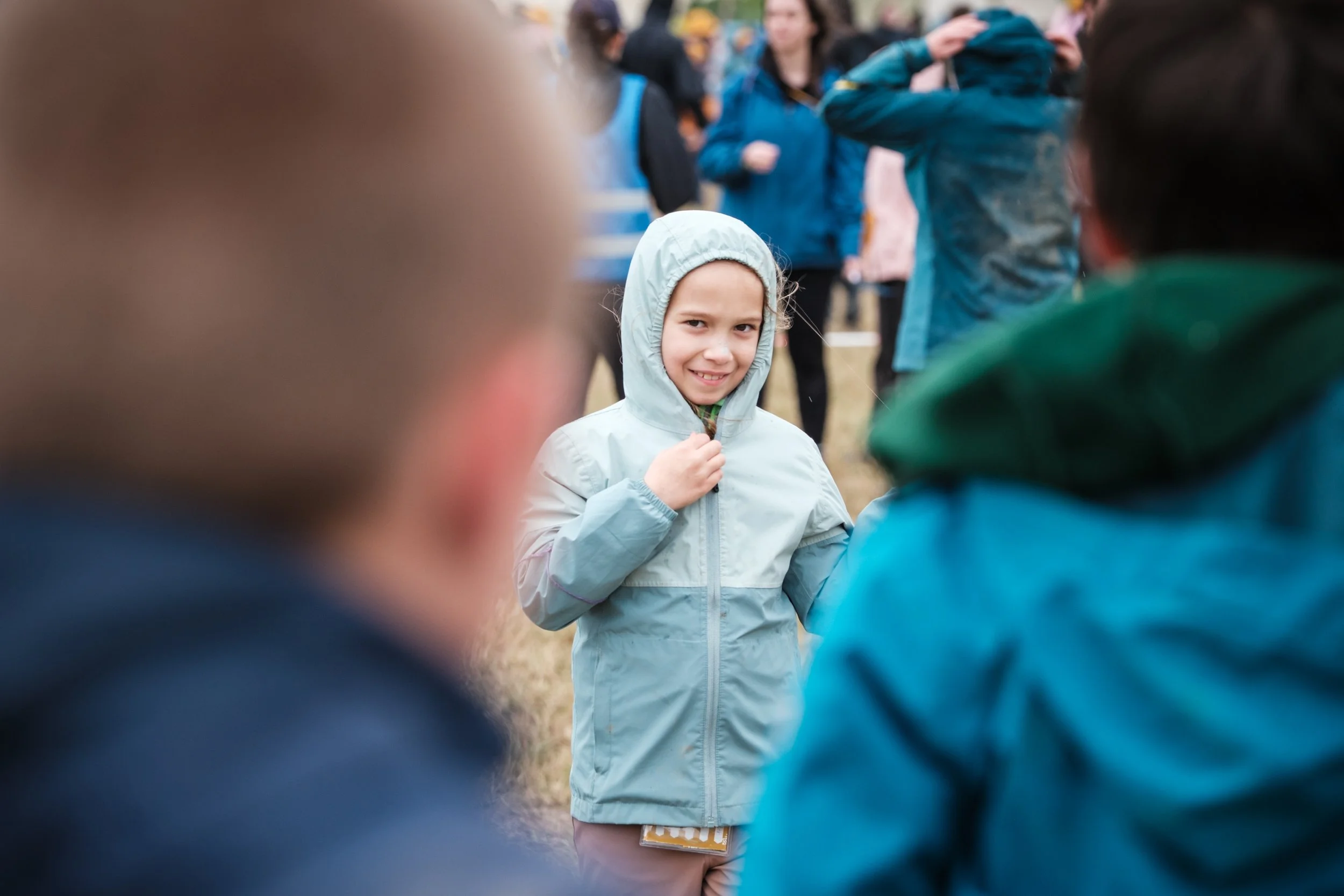 Ein Junge trägt eine hellgraue Regenjacke mit Kapuze und steht im Mittelpunkt, während Menschen im Hintergrund und im Vordergrund um ihn herumgehen, wahrscheinlich bei einem Outdoor-Event.