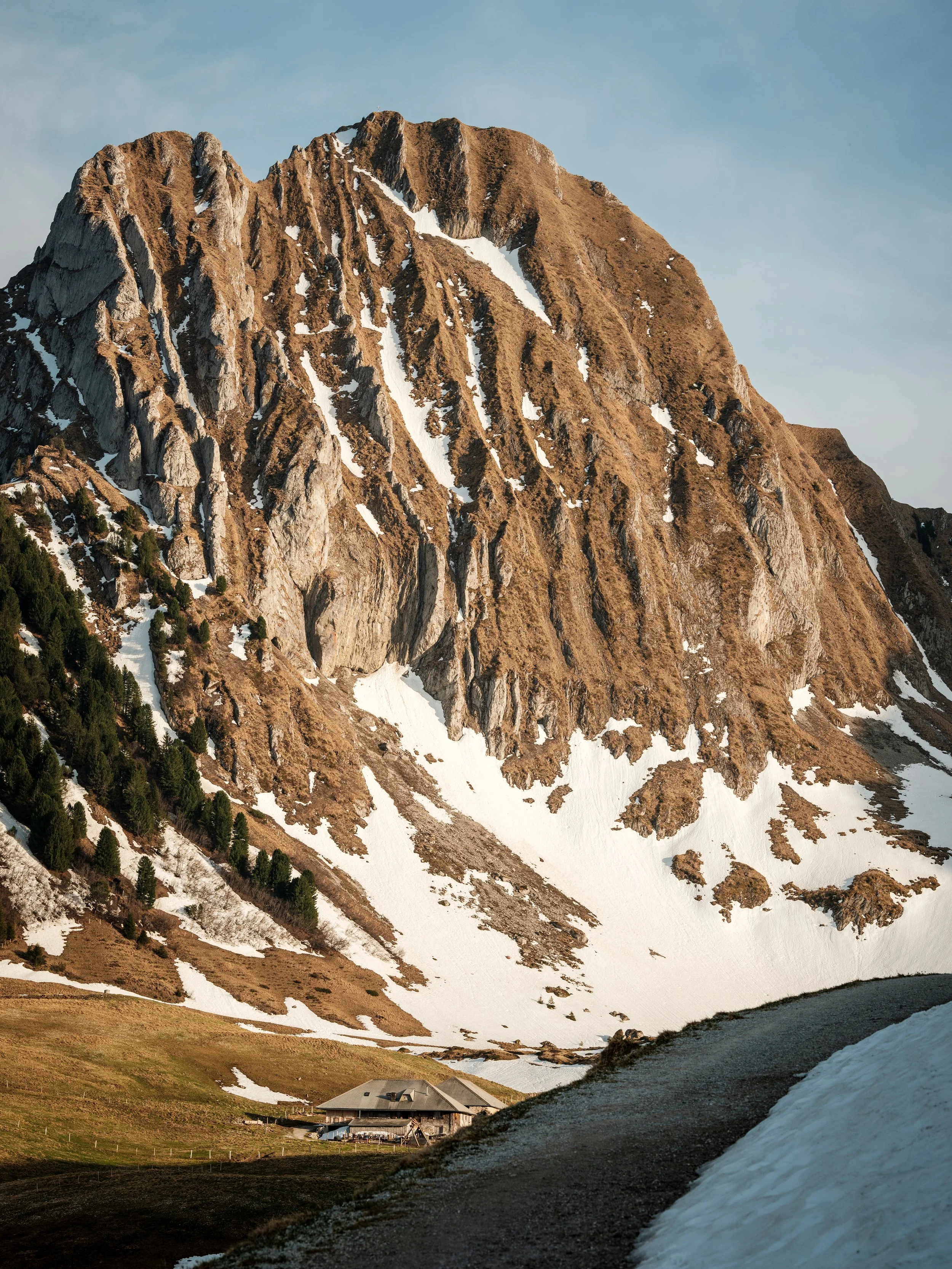 Berg mit steilen Felswänden, teilweise schneebedeckt, mit einem kleinen Haus im Tal.