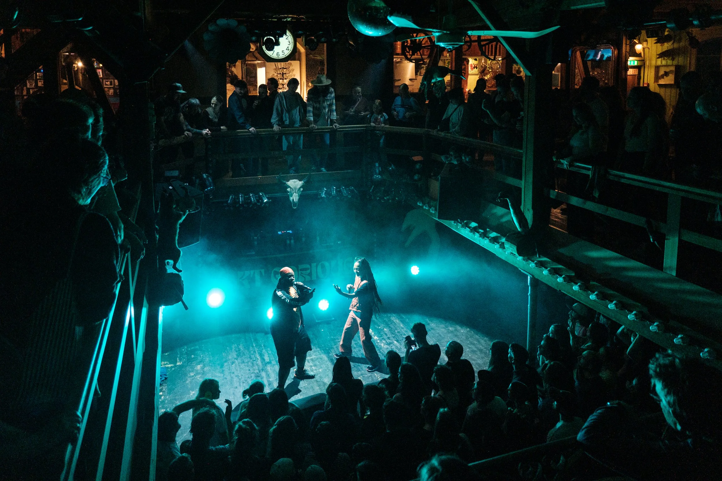 Zwei Personen tanzen auf einer Bühne in einer Bar oder einem Club, im Hintergrund leuchtende blaue Lichter, umgeben von einem Publikum, das zuschaut.