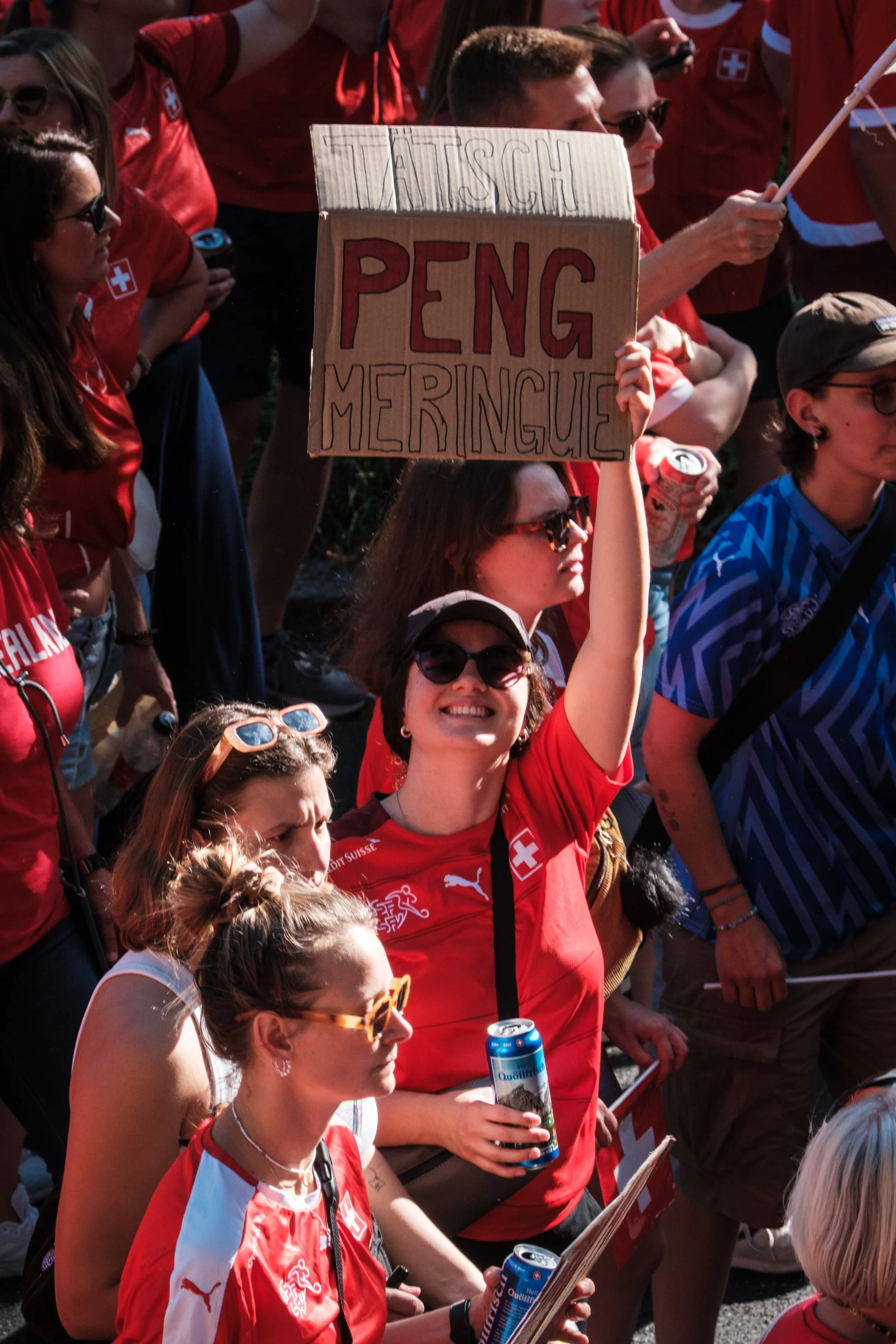 Eine Gruppe von Menschen trägt rote Schweizer Fahnen- und Teamtrikots bei einer Demonstration oder einem Straßenfest. Eine Frau hält ein Schild hoch, auf dem in großen Buchstaben "TASCH PENG MERINGUE" steht. Viele Menschen tragen Sonnenbrillen und ha
