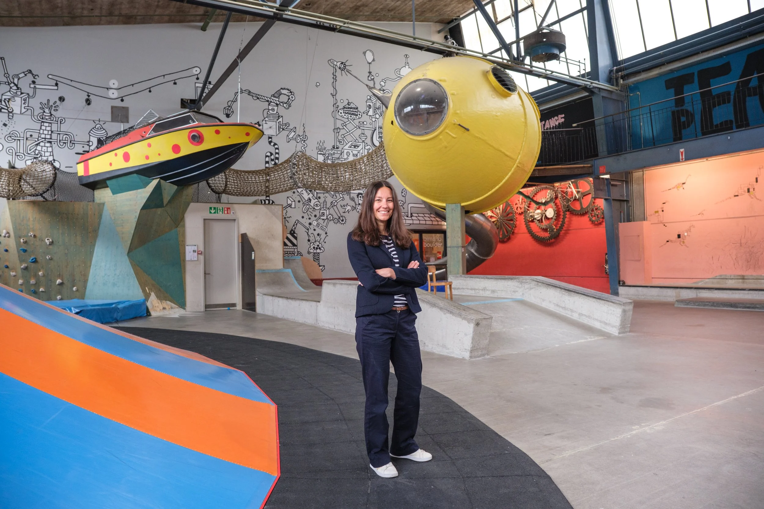 Eine junge Frau in dunkler Kleidung steht in einer Indoor-Kletterhalle mit bunten Kletterwänden, einer Rutsche, einer großen gelben kubusförmigen Struktur und Wanddekorationen mit Zahnrädern und Maschinenteilen.
