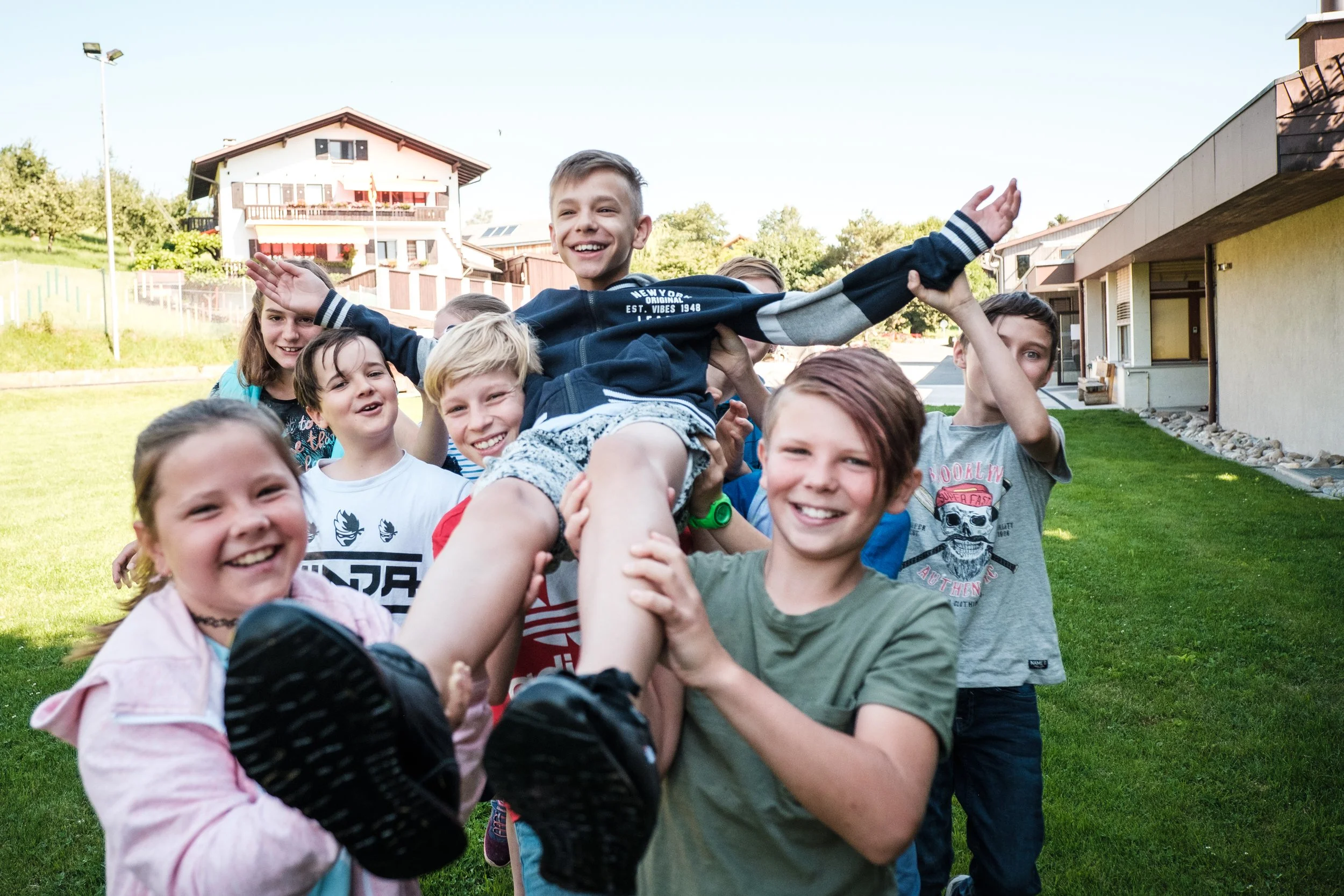 Jugendliche festhalten einen lachenden Jungen, der in die Luft gehoben wird, auf einer grünen Rasenfläche vor einem Haus.