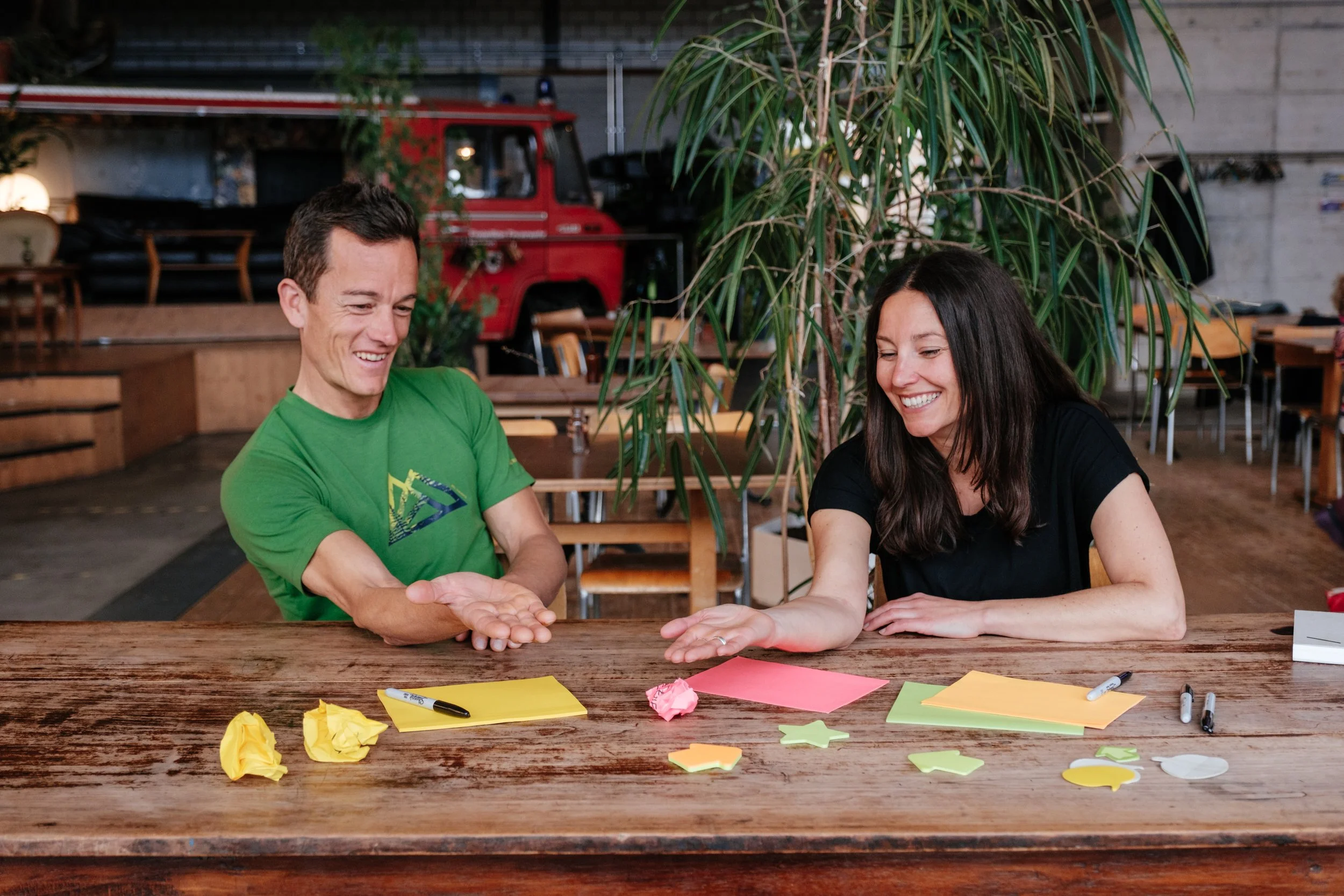 Zwei Menschen sitzen an einem Holztisch, lachen und tauschen Gegenstände aus, während sie Papierausschnitte, Marker und Notizzettel auf dem Tisch haben, in einem Raum mit Holz- und Pflanzenmöbeln.