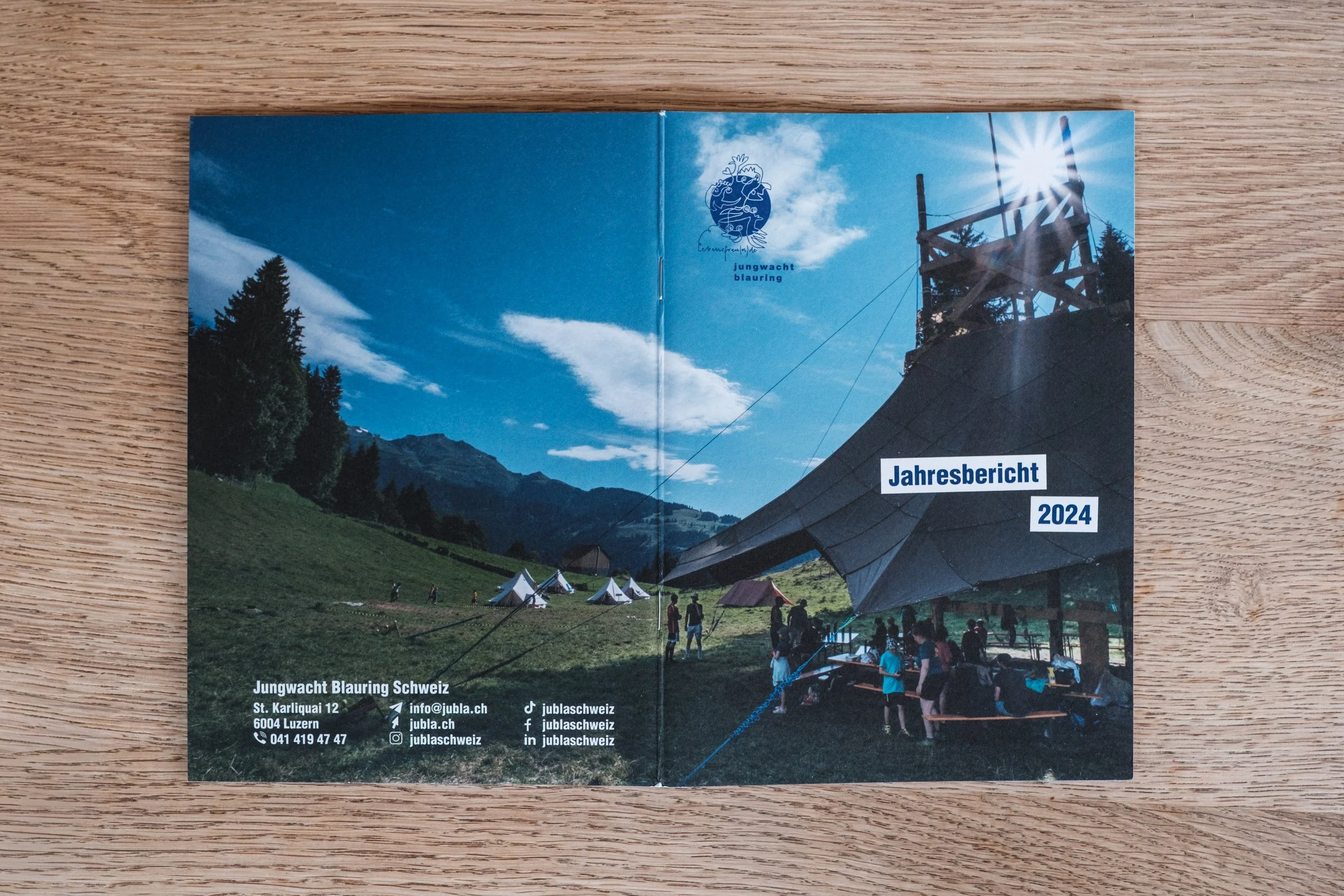 Titelseite des Jahresberichts 2024 von Jungwacht Blauring Schweiz, mit einem Bild von einem Zeltlager in den Bergen, Zelten, Gruppen von Menschen und einem großen Pavillon, blauem Himmel und Bergen im Hintergrund.
