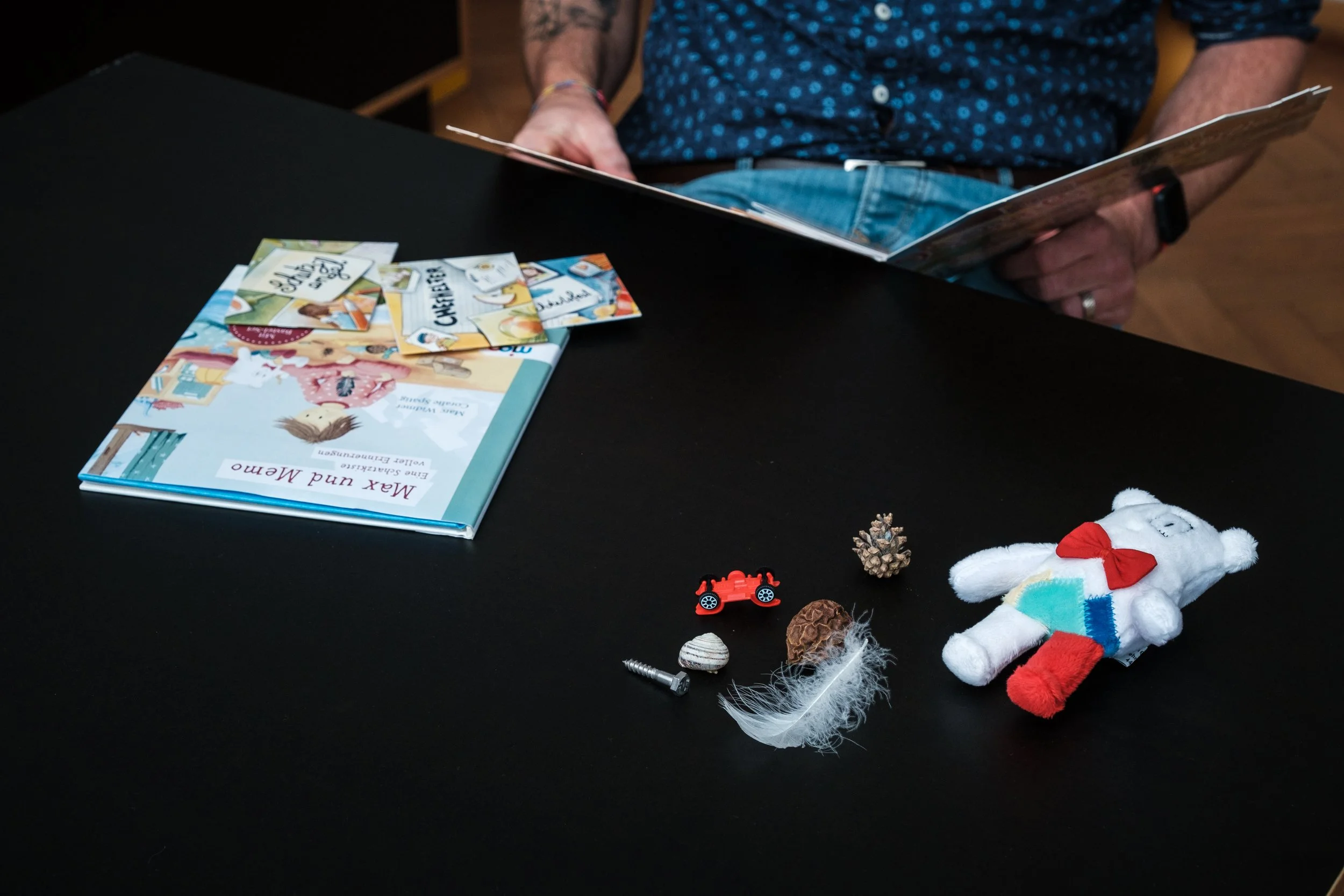 Offener Bilderbuch auf einem schwarzen Tisch mit verschiedenen Spielzeugen und Gegenständen, darunter ein Stofftier, Stift, Schraube, kleine Naturgegenstände und eine Miniaturauto.