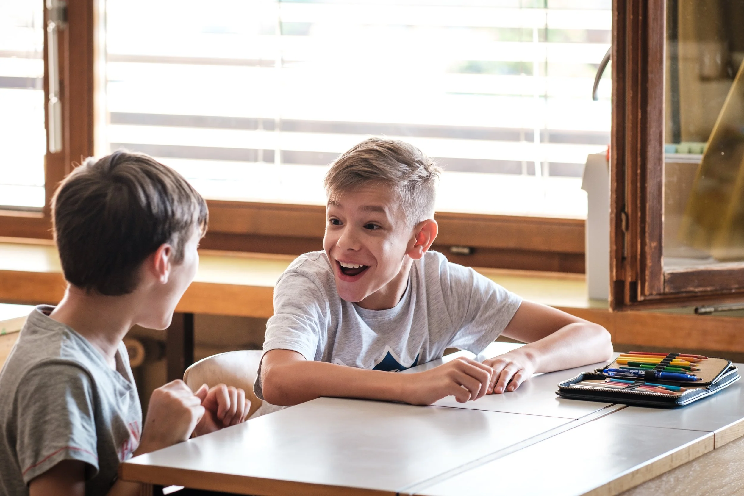 Zwei Jungen sitzen sich gegenübersitzend an einem Tisch, lachend und beim Gespräch, mit Buntstiften und Filzstiften vor ihnen auf dem Tisch.
