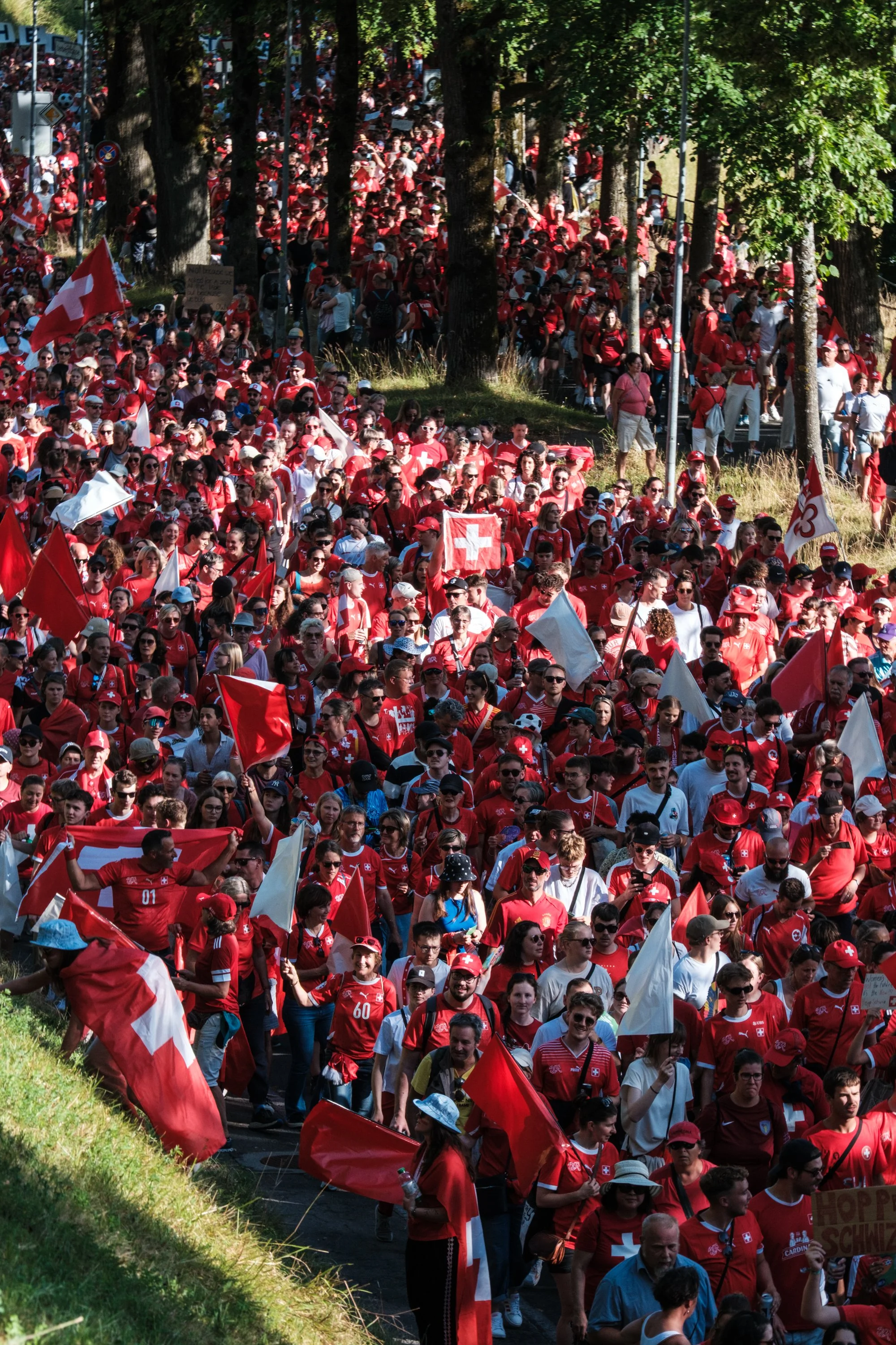Menschenmenge bei einem Schweizer Fussball-Fanmarsch, alle in roten Trikots und mit Flaggen. Im Hintergrund Bäume und mehr Leute, die den Weg entlang gehen.