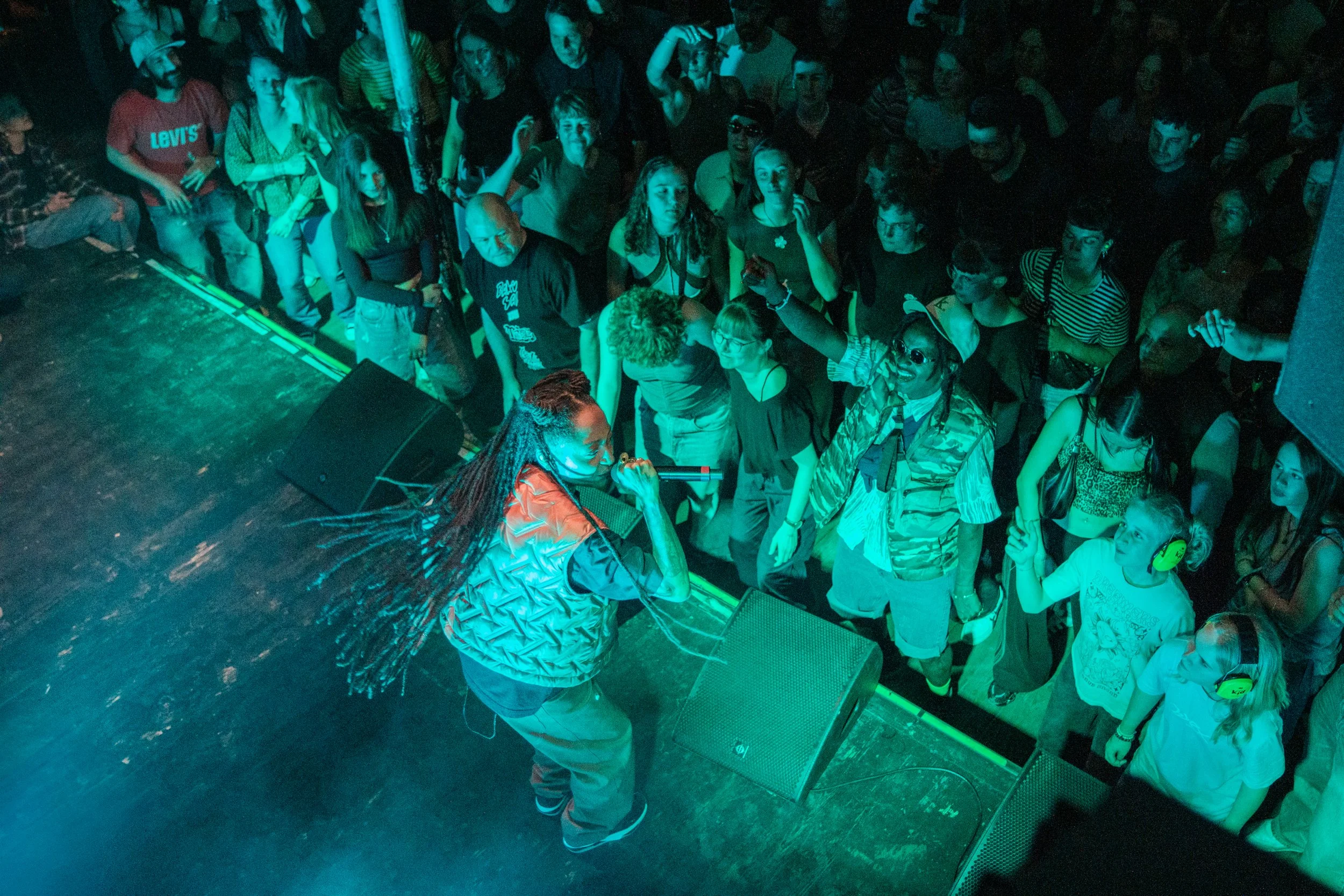 Ein Performer mit langen Zöpfen singt auf der Bühne vor einem Publikum in einem dunklen Raum bei einer Live-Performance.