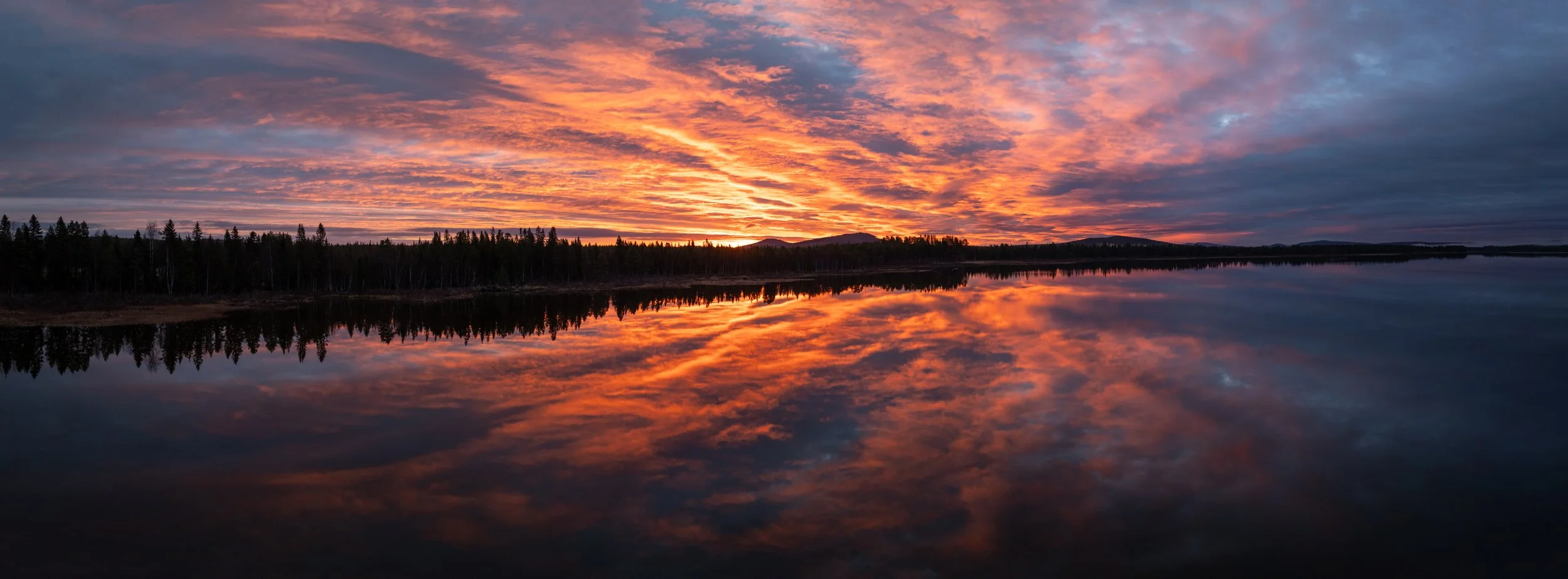 Sonnenuntergang über einem ruhigen See mit bewölktem Himmel und reflektierendem Wasser, umgeben von einem Wald im Hintergrund.