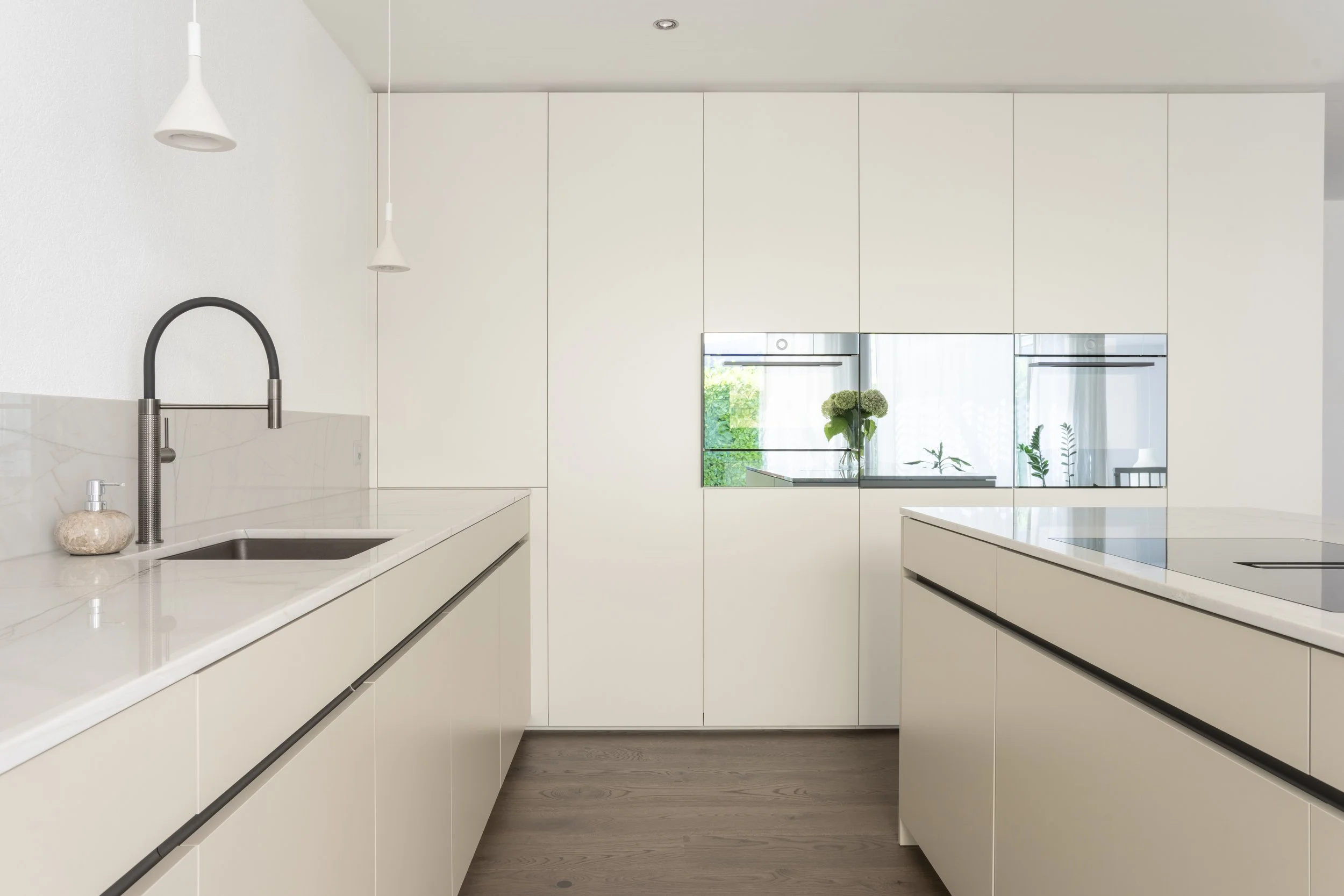Moderne, minimalistische Küche mit weißen Schränken, einer schwarzen Spüle mit schwarzem Wasserhahn, Holzfußboden und einem Fenster mit Blick auf grüne Pflanzen.