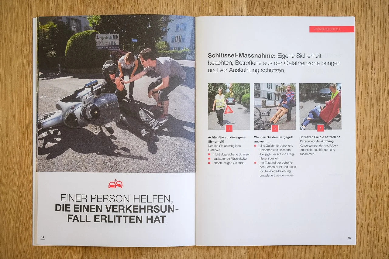 Offenbar in einer Broschüre über Verkehrssicherheit, zeigt eine Szene, in der eine Person nach einem Verkehrsunfall von mehreren Helfern auf dem Boden betreut wird. Links im Bild ist eine verunglückte Motorrad auf der Straße zu sehen.