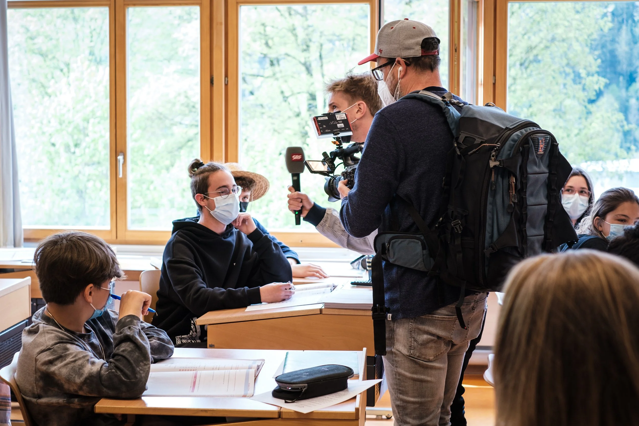Ein Lehrer und mehrere Schüler in einem Klassenzimmer, alle tragen Masken. Der Lehrer filmt die Schüler mit einer Kamera, während diese am Unterricht teilnehmen.
