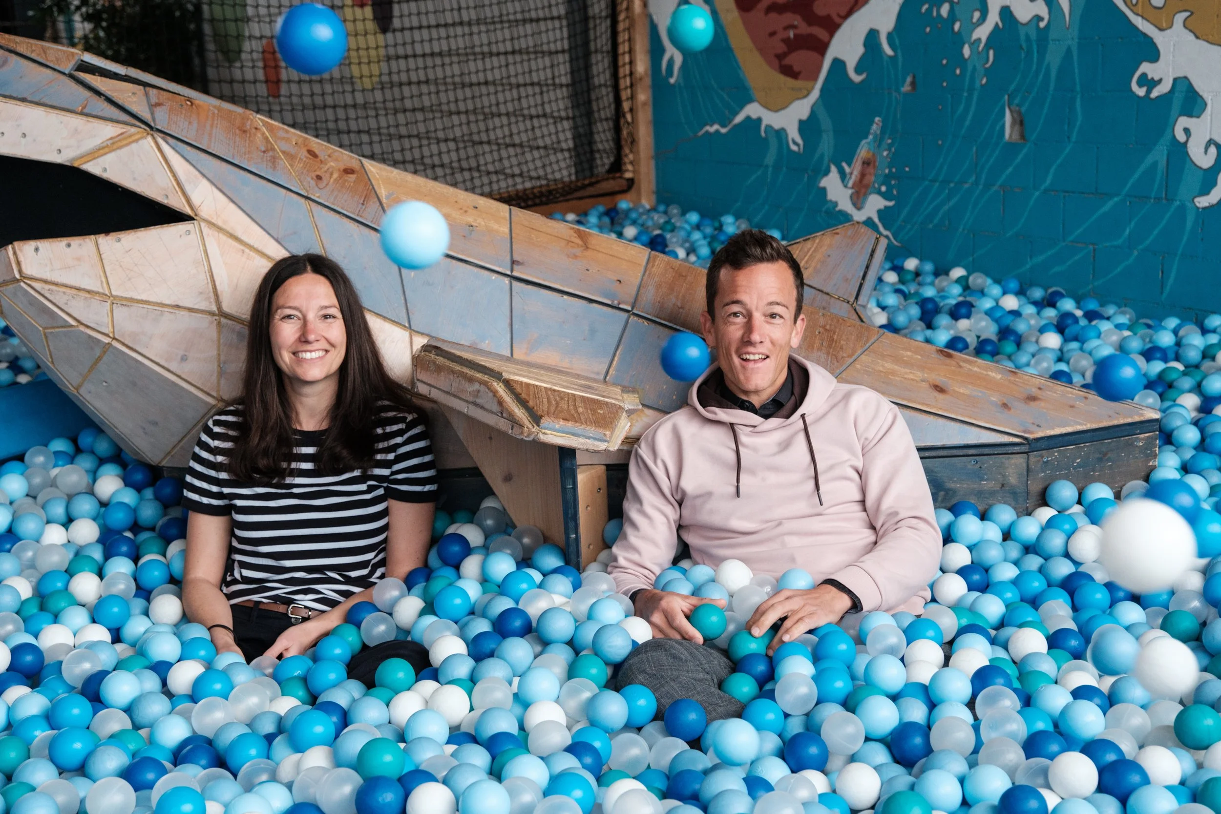 Zwei lächelnde Menschen sitzen in einer Ballgrube mit blauen, weißen und hellblauen Bällen, hinter ihnen ein Klettergerüst und eine Wand mit Wandkunst.