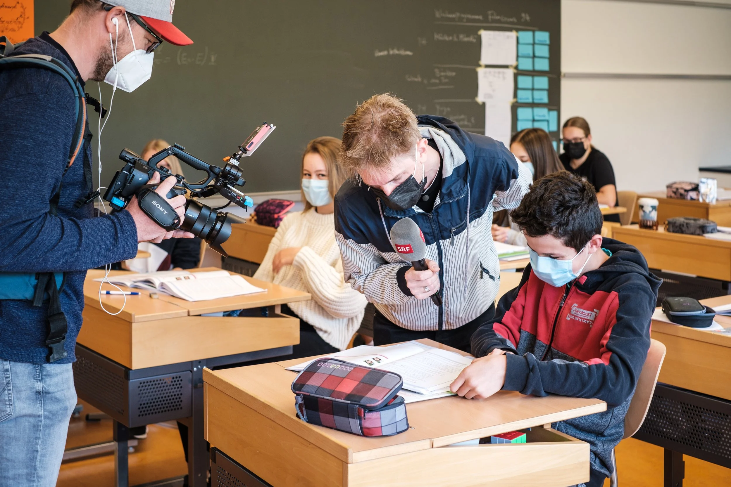 Ein Lehrer interviewt einen Schüler in einer Klasse während eines Filmdrehs. Der Lehrer hält ein Mikrophon, der Schüler trägt eine Maske, liest ein Buch, und im Hintergrund sitzen weitere Schüler in einer Schulklasse.