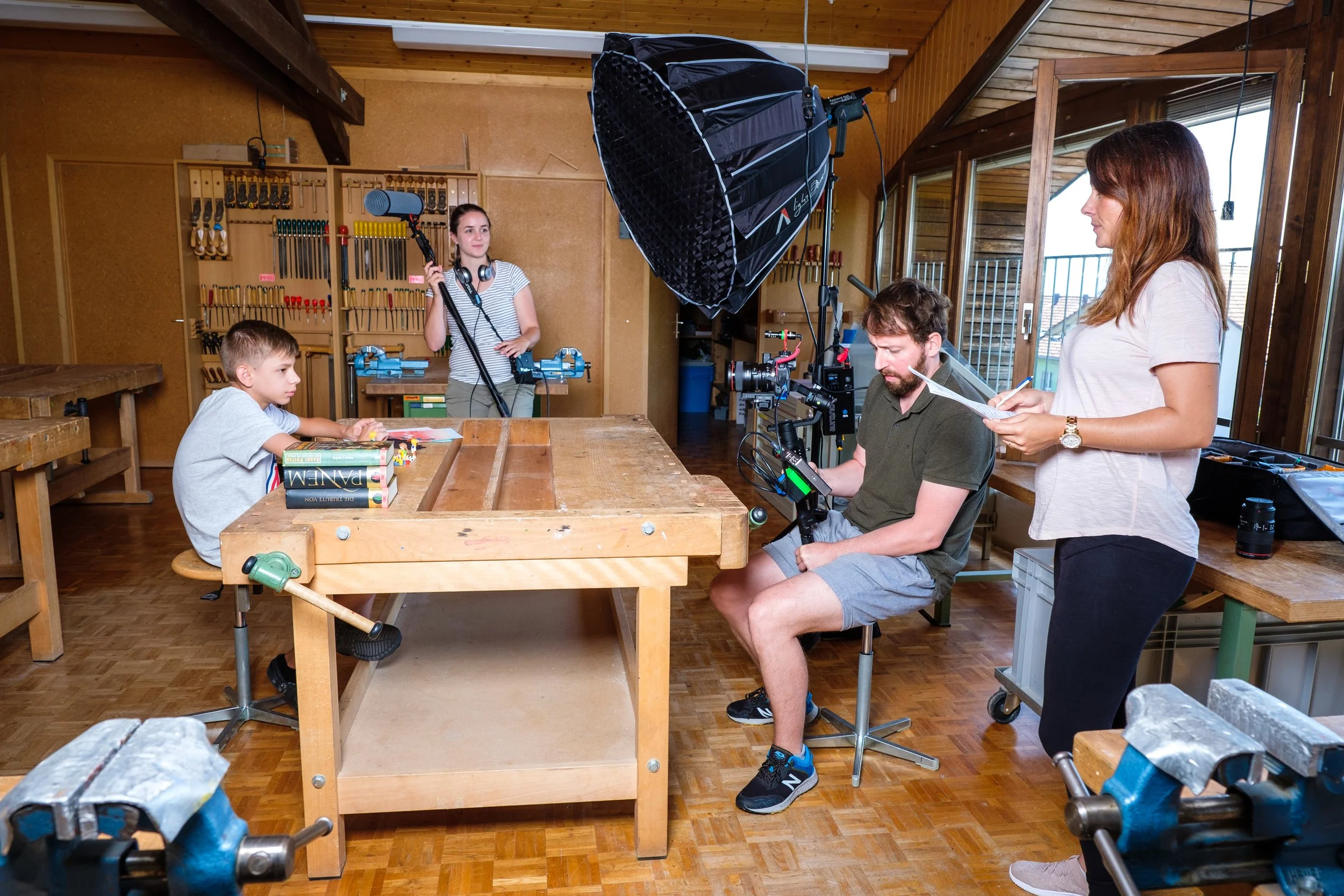 Ein Set für einen Film- oder Videodreh in einer Holzwerkstatt. Es gibt eine junge Frau mit einem Mikrofon und Kopfhörern, einen jungen Mann, der an einer Kamera sitzt, eine Frau mit Notizen und ein Kind, das auf einem Werkbank sitzt.