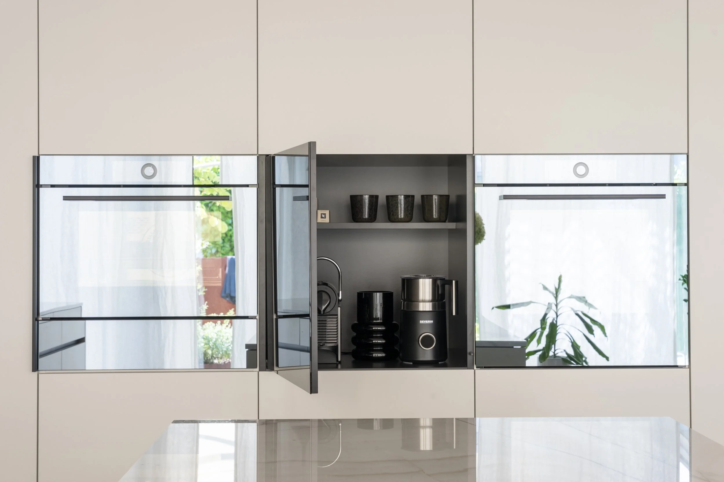 Offene Küchenschrank mit schwarzen Küchengeräten und Dekogefäßen, hinter Glasschrank mit weißen Vorhängen und Garten im Hintergrund.