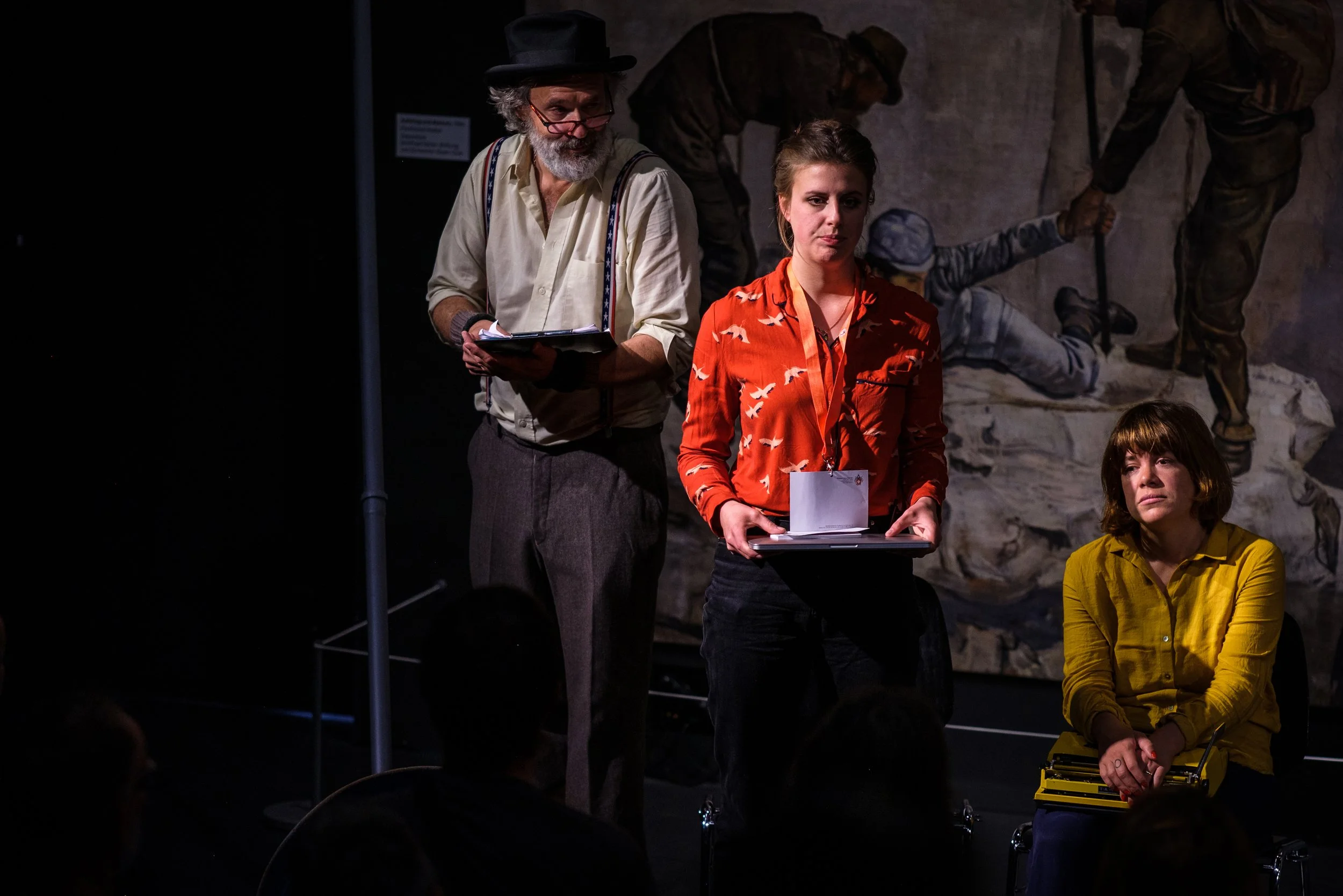 Menschen bei einer Theaterprobe, eine Person liest, eine andere hält Papiere, eine sitzt auf einem Stuhl, im Hintergrund Wandbild.