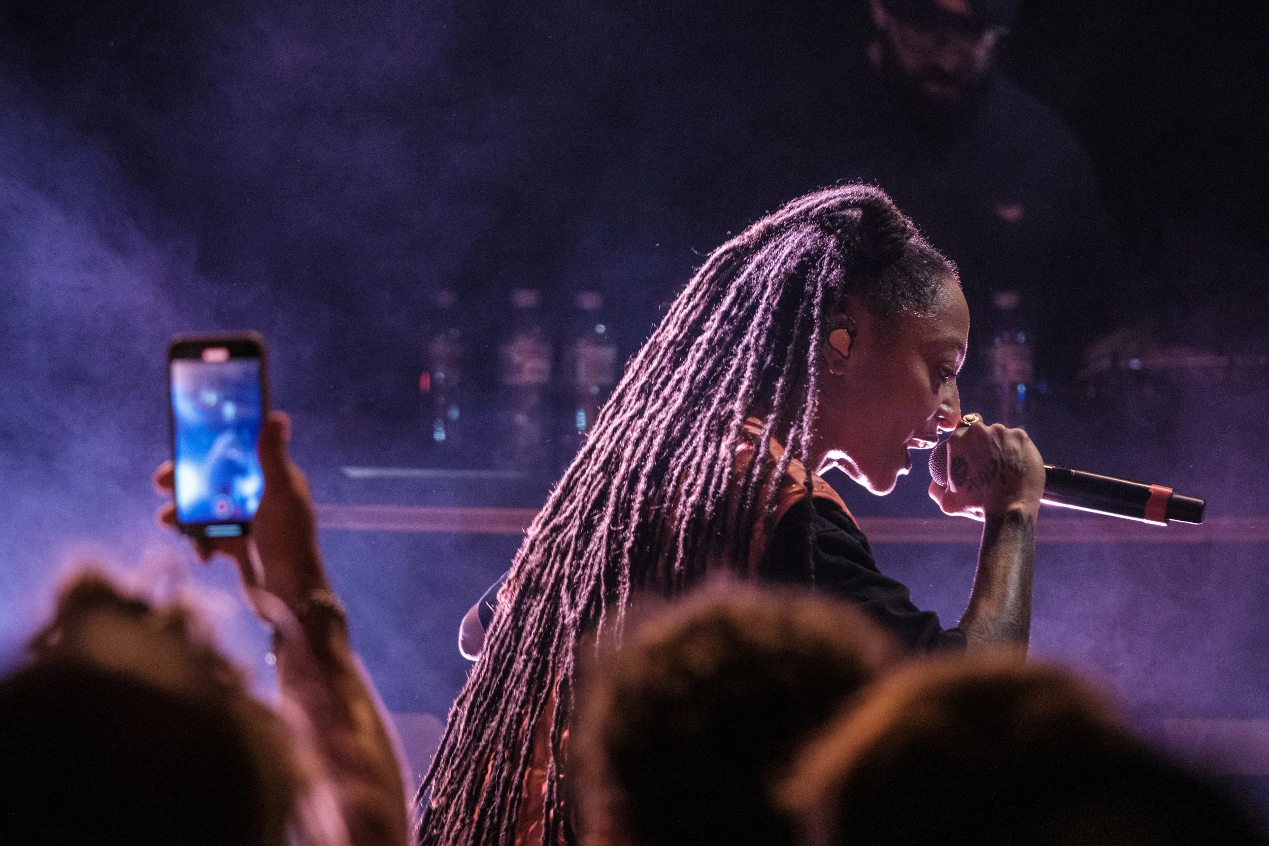 Eine Sängerin mit dreadlocks singt in einem dunklen Raum auf einer Bühne und hält ein Mikrofon. Im Vordergrund ist eine Person, die ein Smartphone benutzt, um die Performance aufzunehmen.
