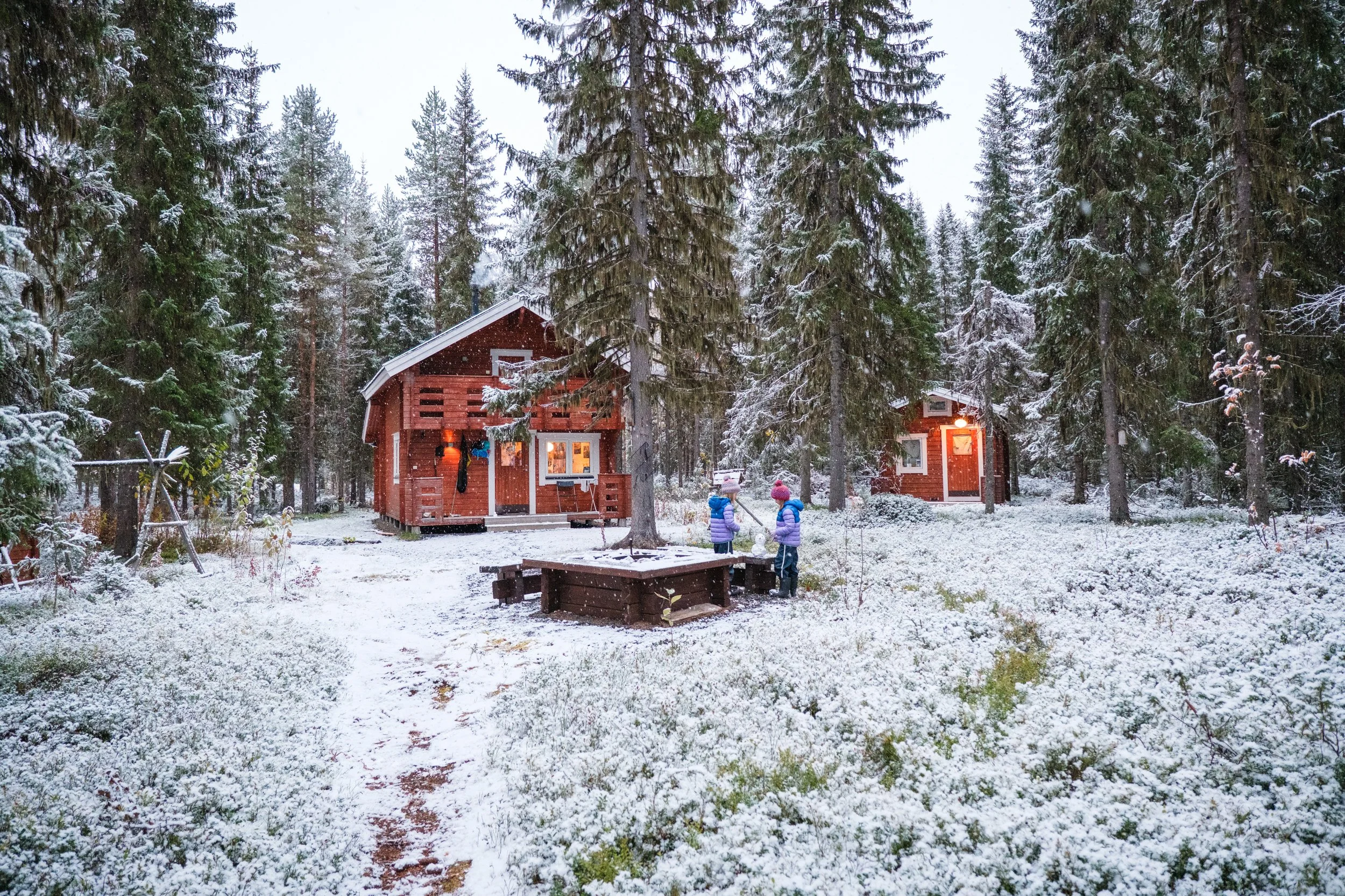 zwei Kinder in Winterkleidung spielen im verschneiten Wald, neben einer kleinen Feuerstelle, vor zwei roten Holzhäusern mit Lichter im Wald